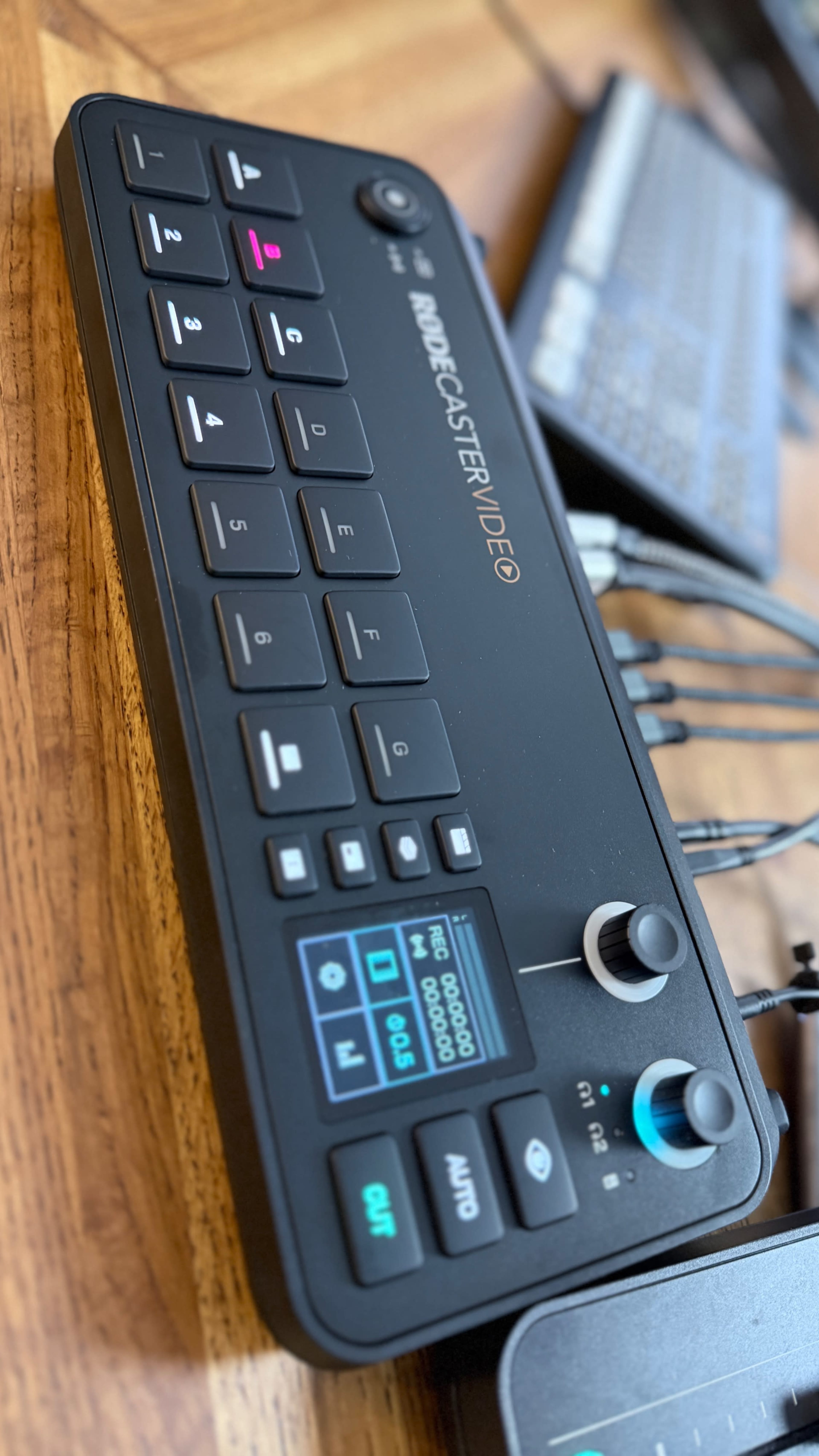 The image shows a close-up of a video and audio control device with various buttons and knobs, resting on a wooden surface beside a keyboard.