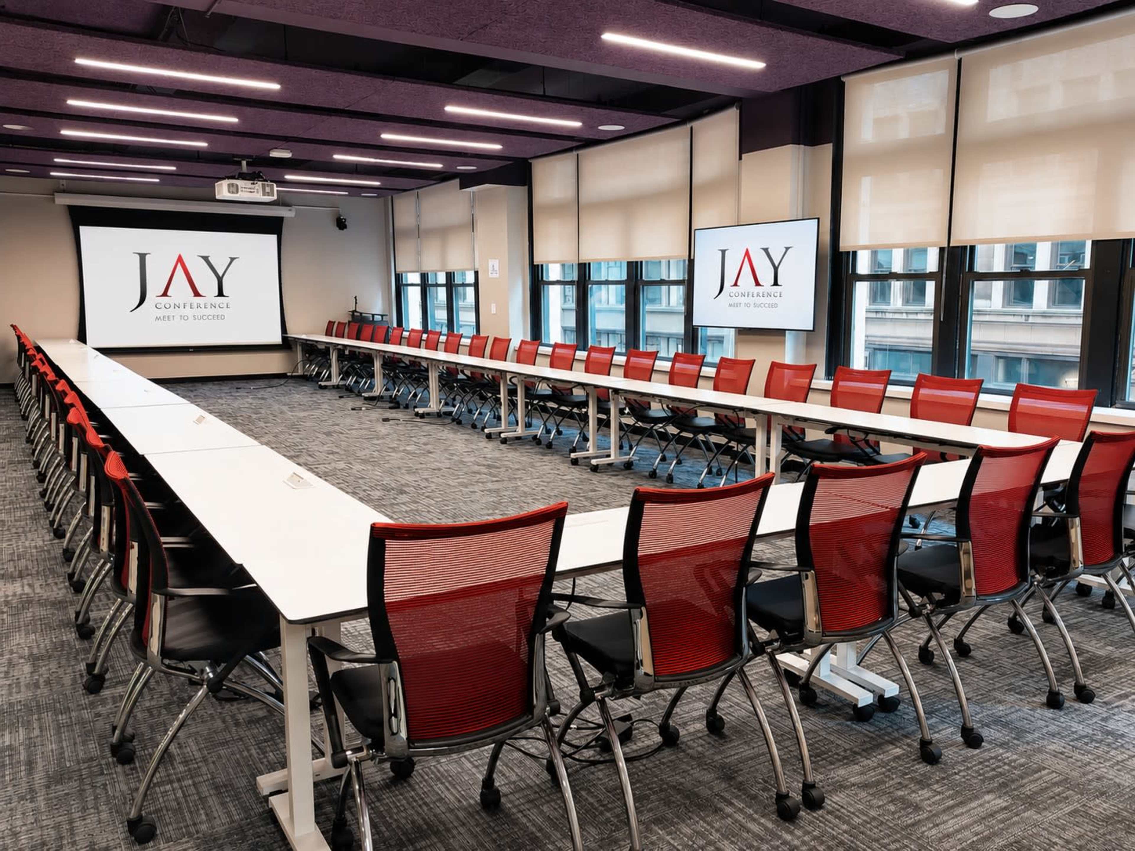 A conference room features a long table surrounded by red and black chairs, with screens displaying the logo "JAY Conference" on the walls.