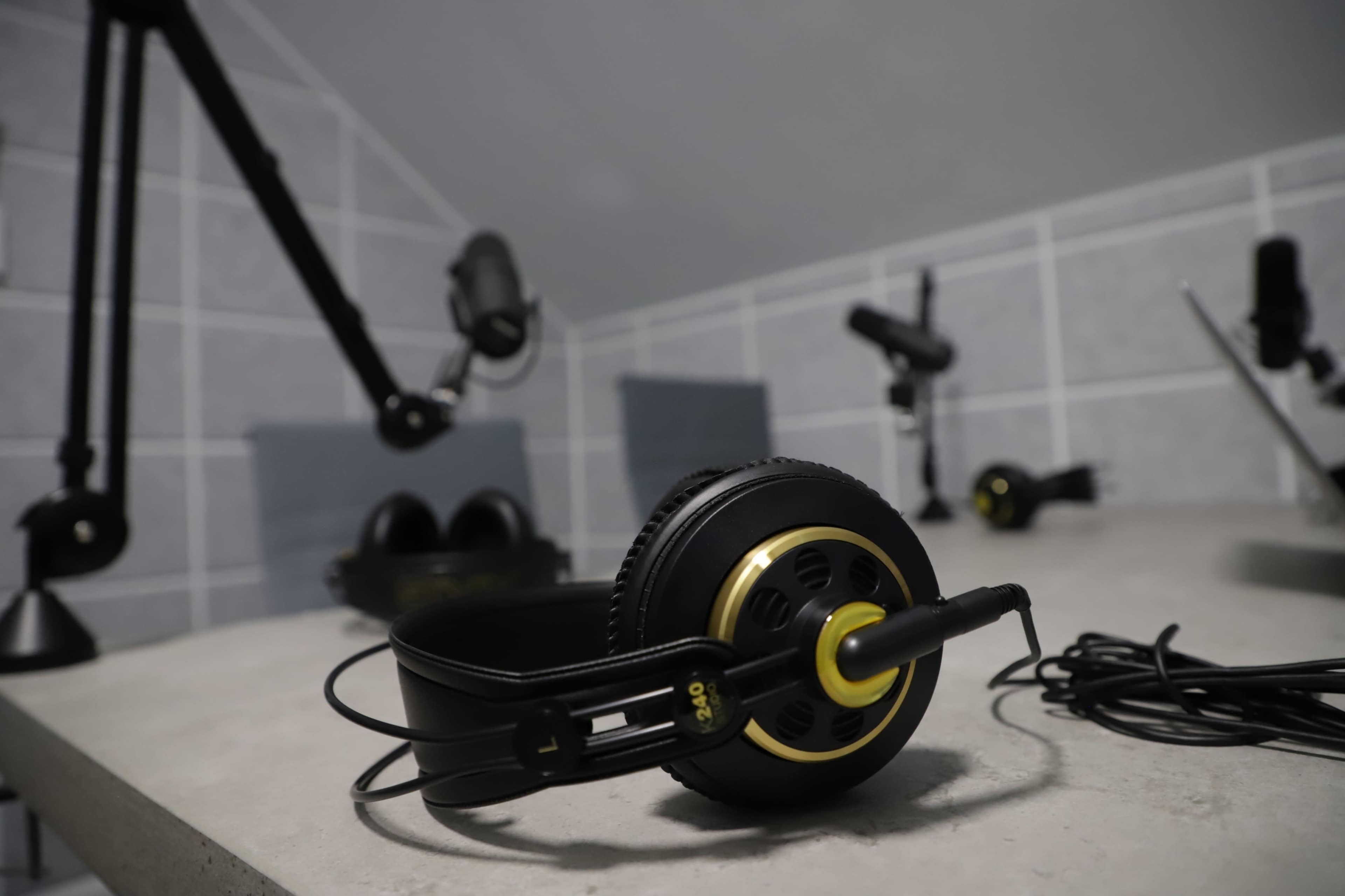 A pair of black headphones with gold accents sits on a concrete surface, surrounded by various microphones and audio equipment in a minimalist workspace.