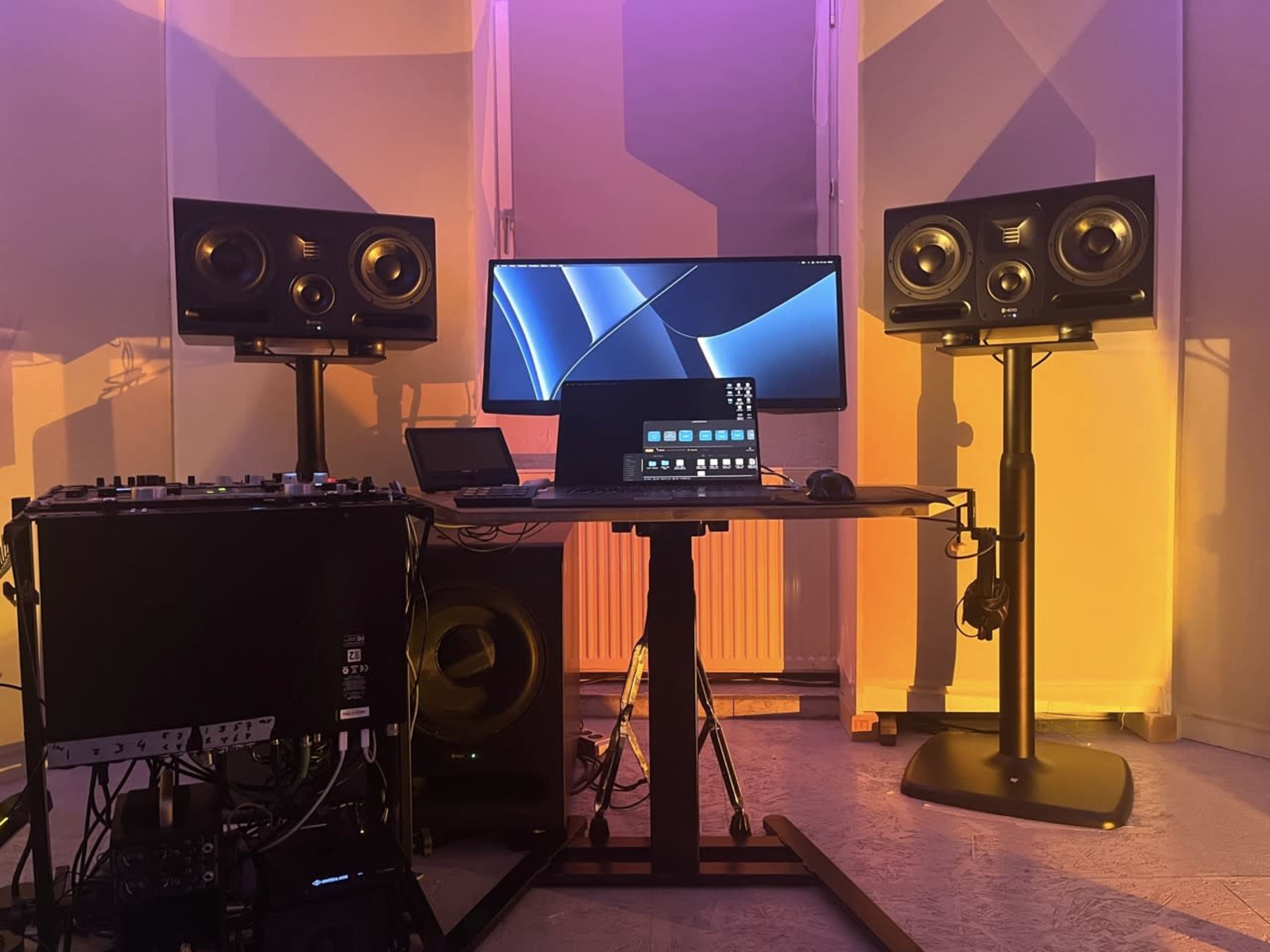 The image shows a recording studio setup with two large speakers, a computer monitor, and various audio equipment arranged on a table in a softly lit room.