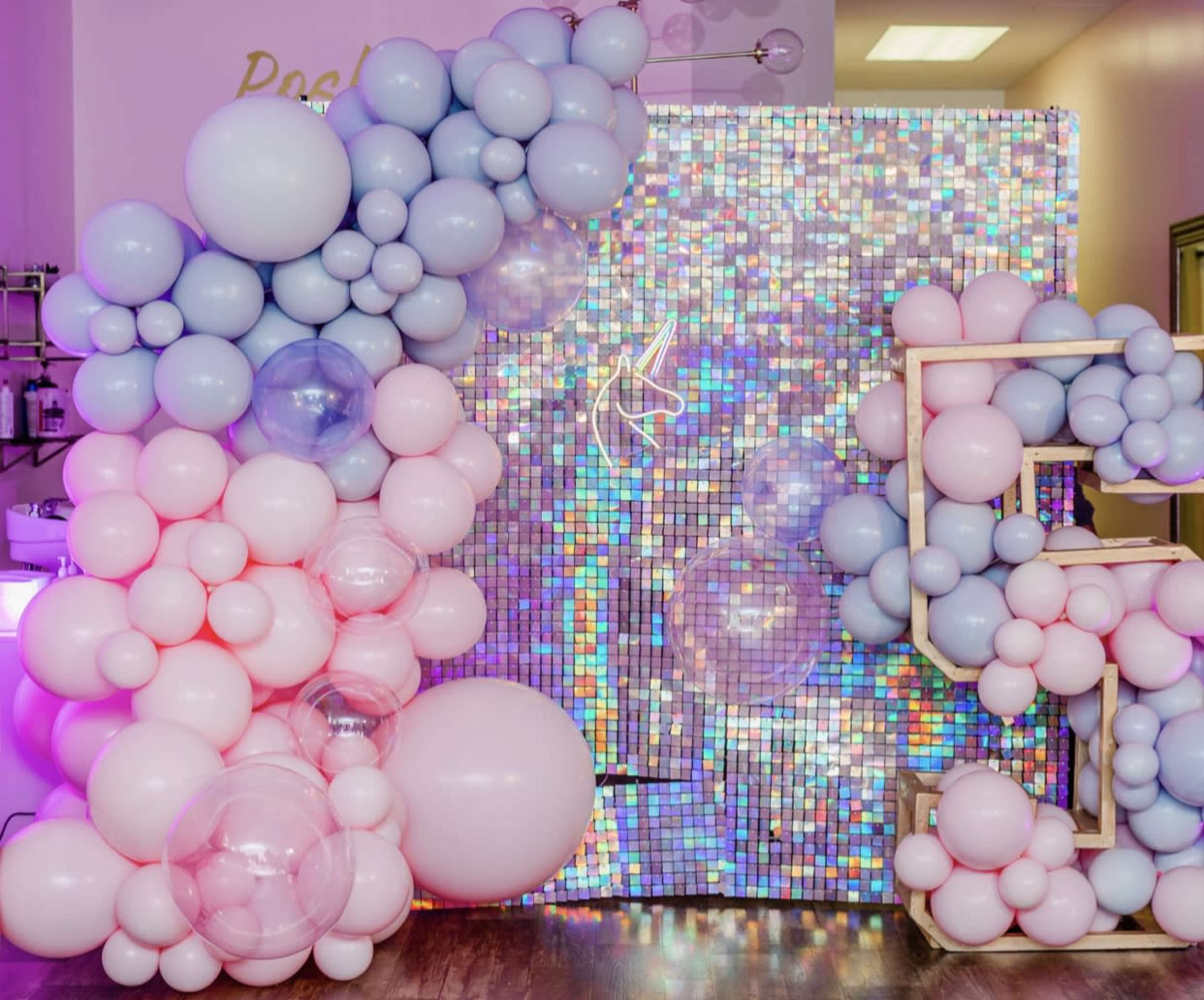 A decorative wall composed of variously sized pink and blue balloons is set against a shimmering, multicolored backdrop.