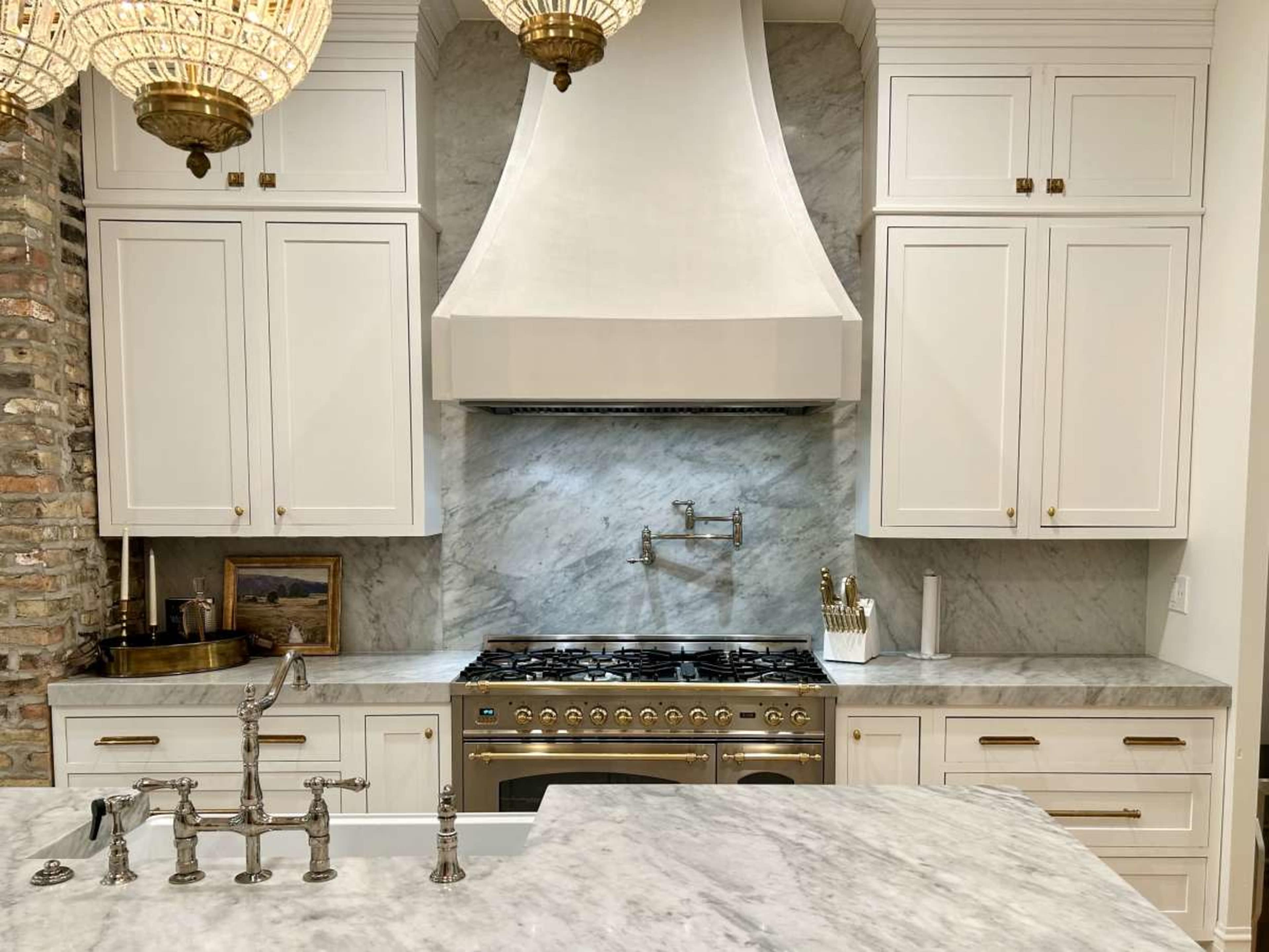 The image shows a modern kitchen with a marble countertop, a large stainless steel range hood, and a professional-grade gas stove beneath it.