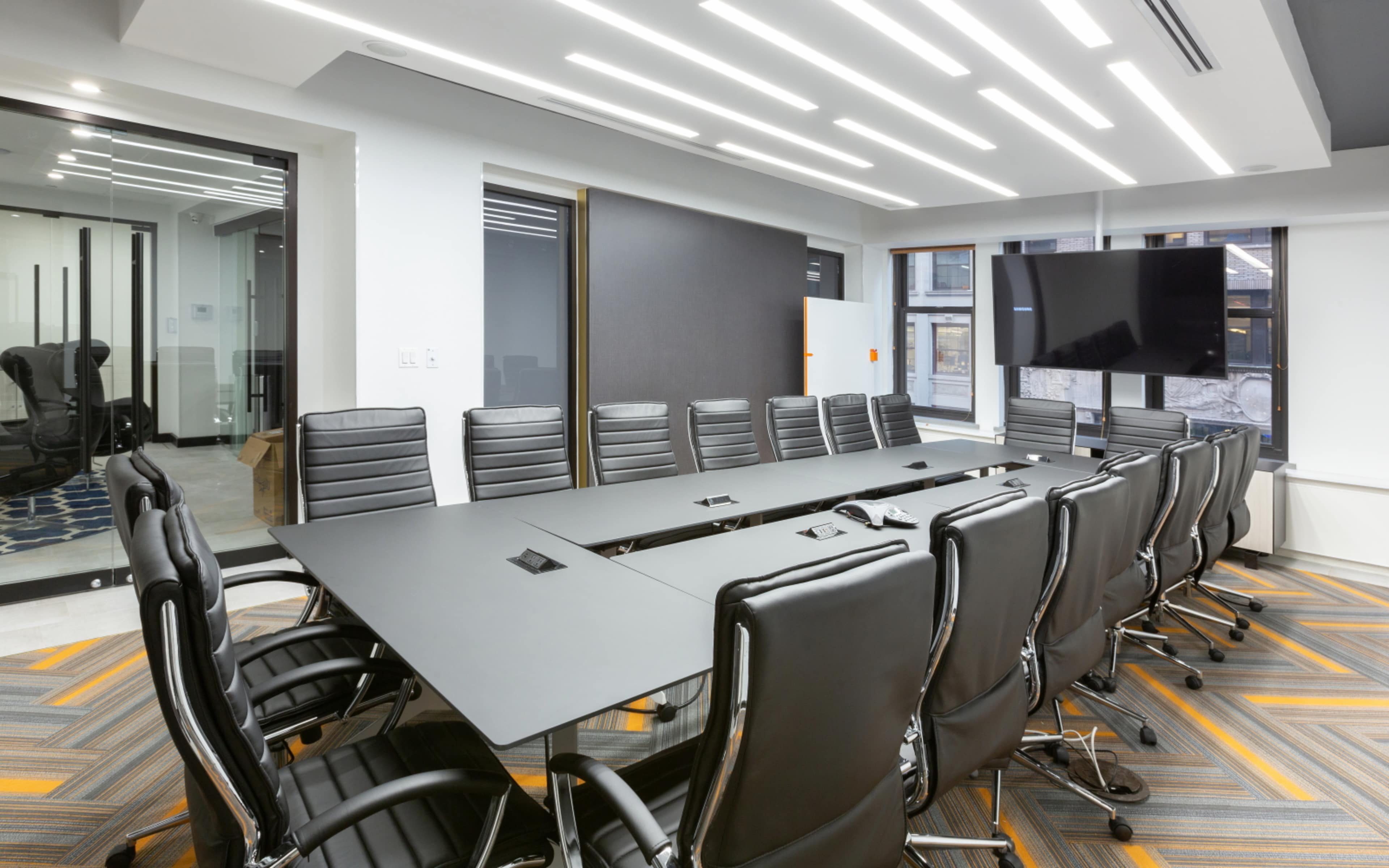 A modern conference room features a long black table surrounded by multiple black ergonomic chairs, with a large television mounted on the wall and illuminated by overhead LED lights.