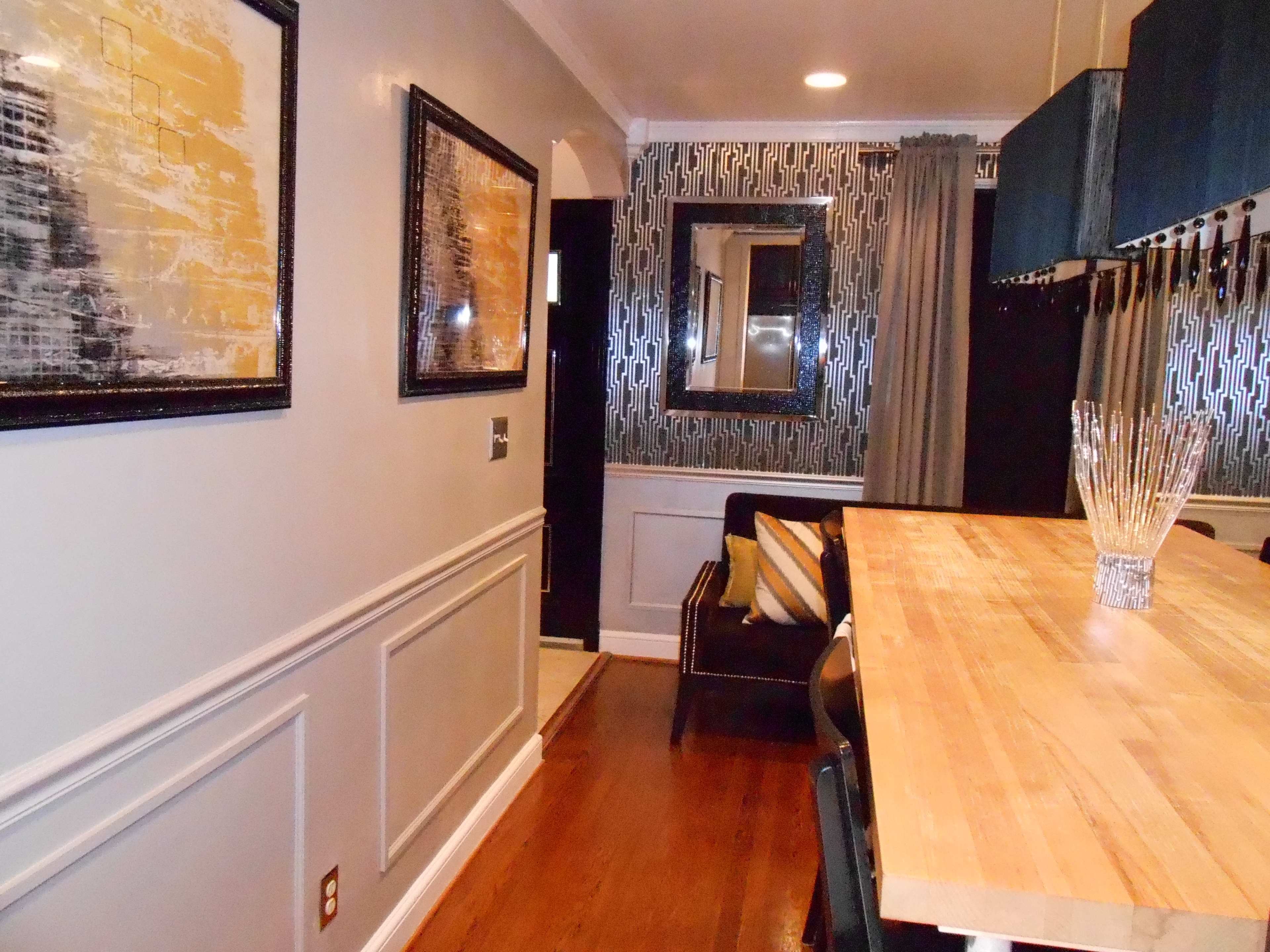 A hallway leading into a dining area with a wooden table, framed artworks on the walls, and a patterned wallpaper in the background.