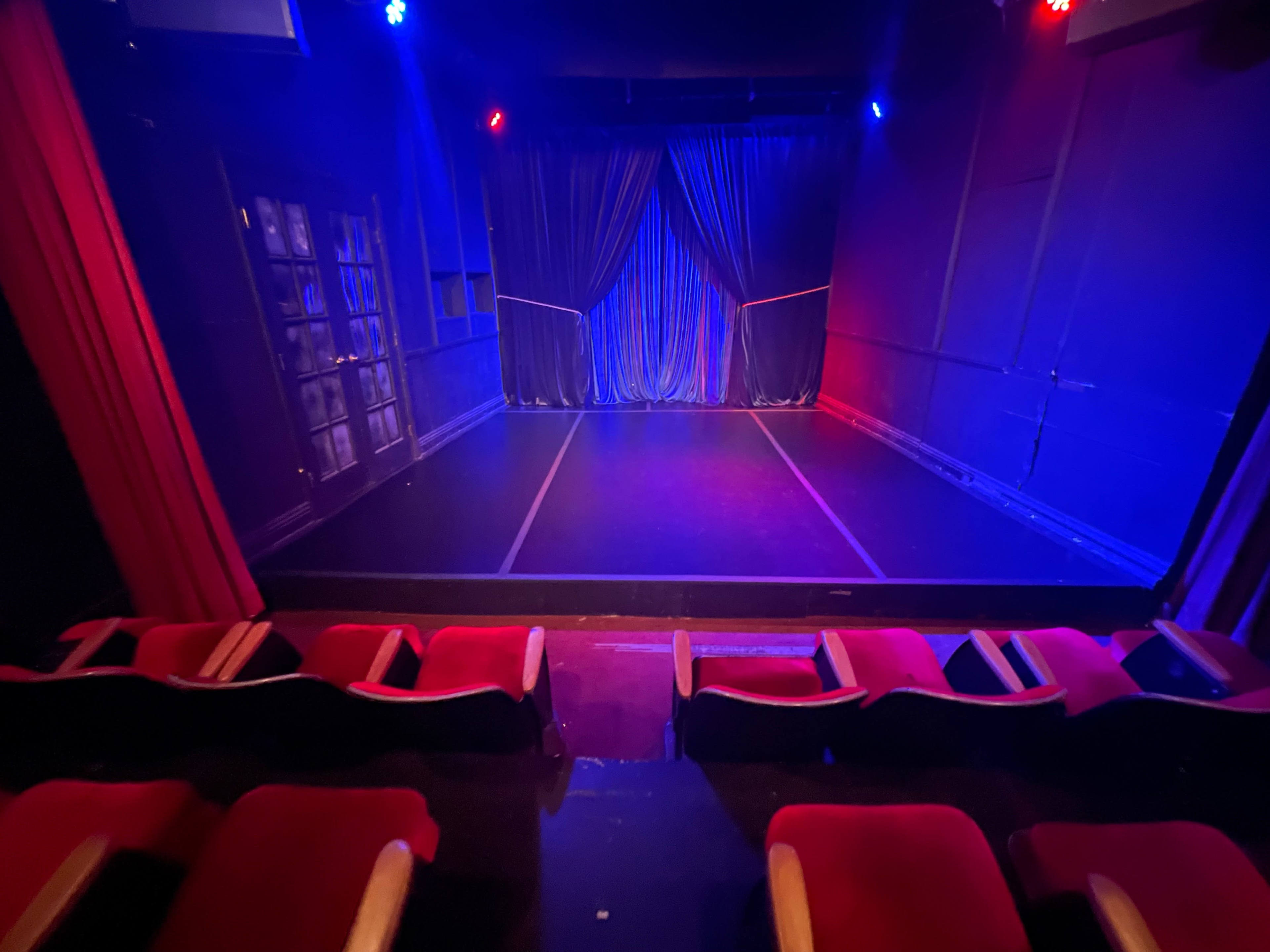 A dark theater stage with red and blue lighting, featuring a curtain in the center and empty red seats in the foreground.