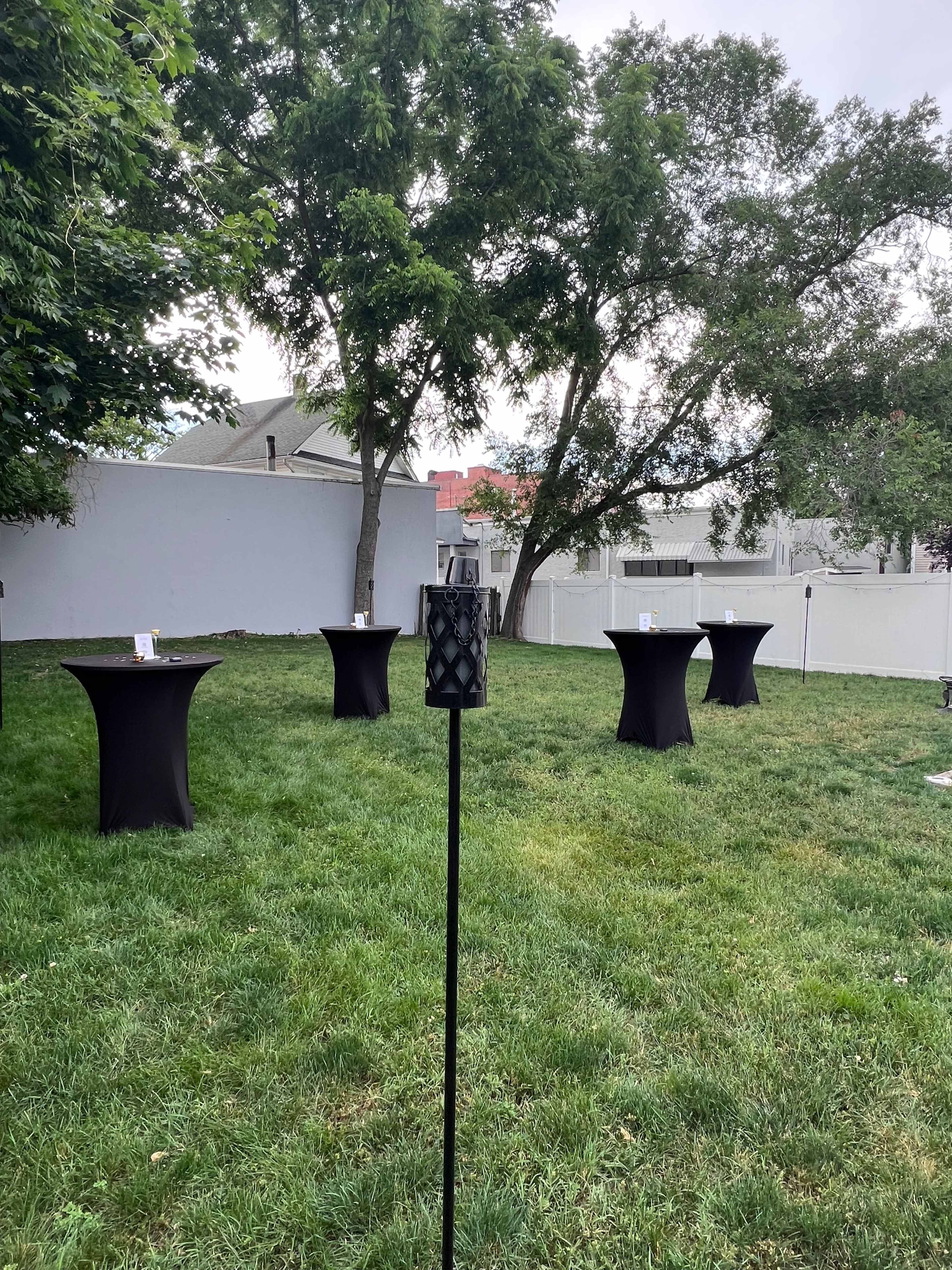 A grassy backyard with several black cocktail tables arranged around a central area, and a decorative torch in the foreground.
