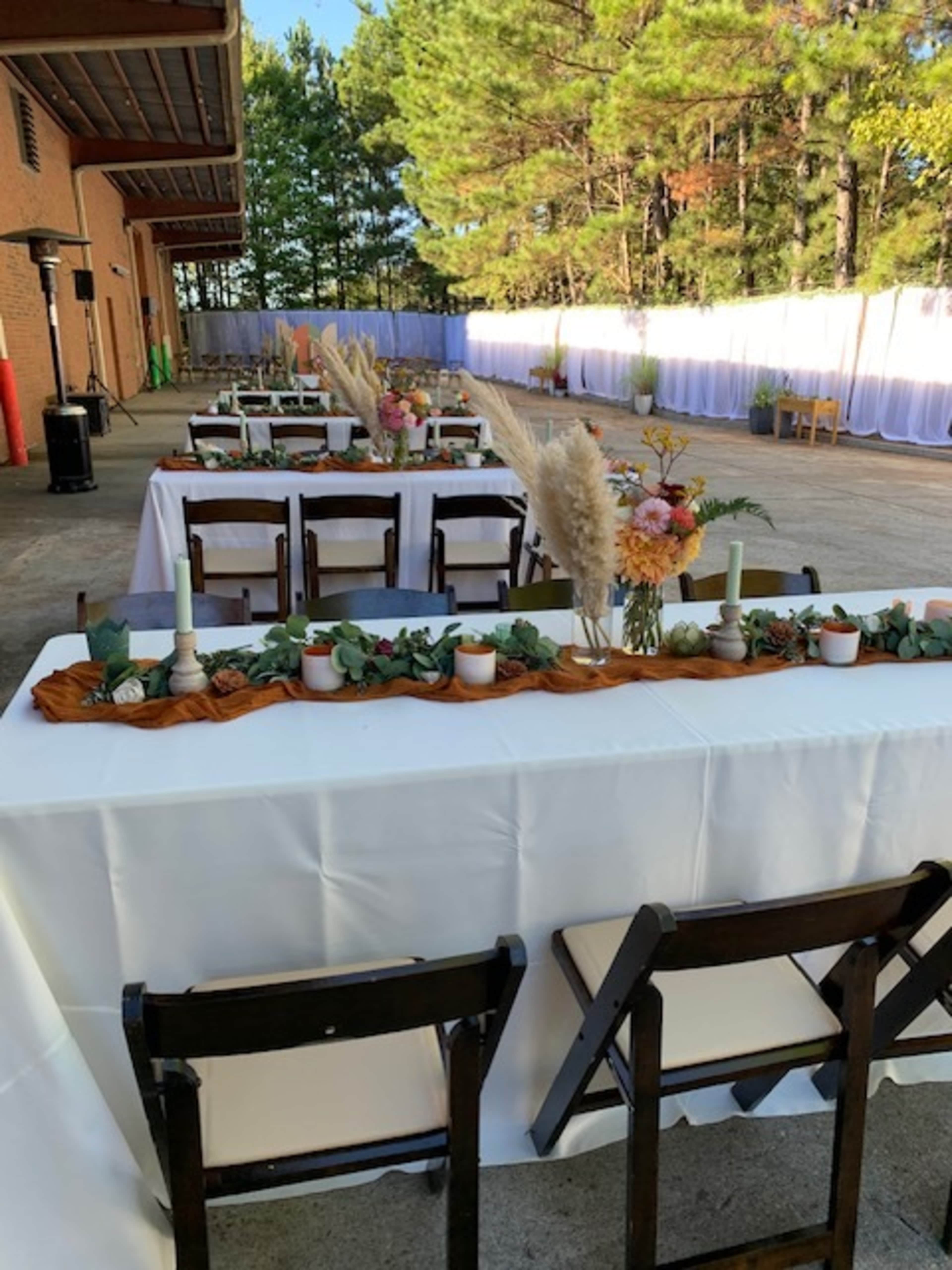 A series of tables are set up outside, decorated with floral arrangements and candles, under a backdrop of trees.