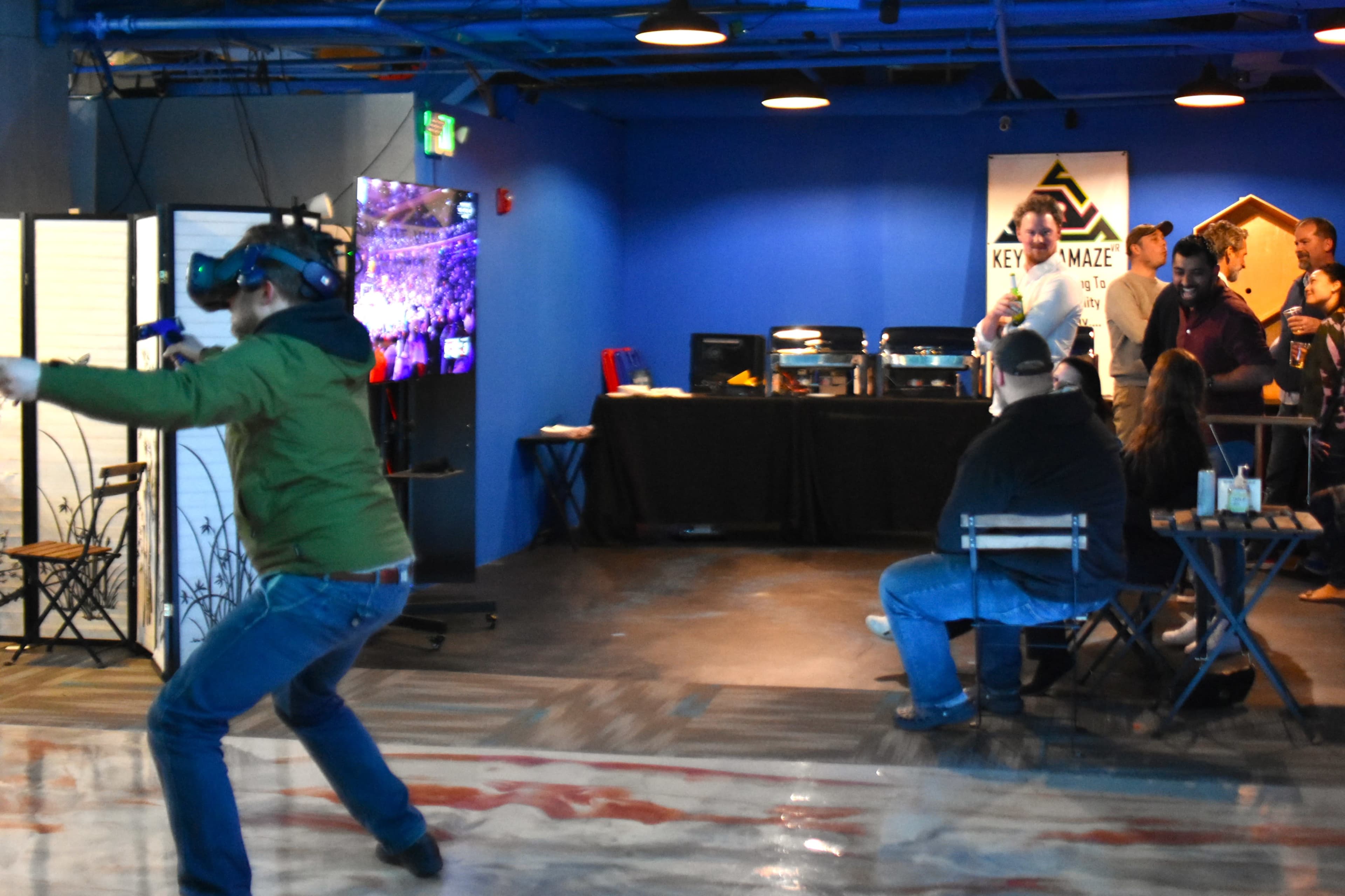 A person wearing a virtual reality headset is engaged in an activity in a colorful arcade space, while others observe and interact in the background.