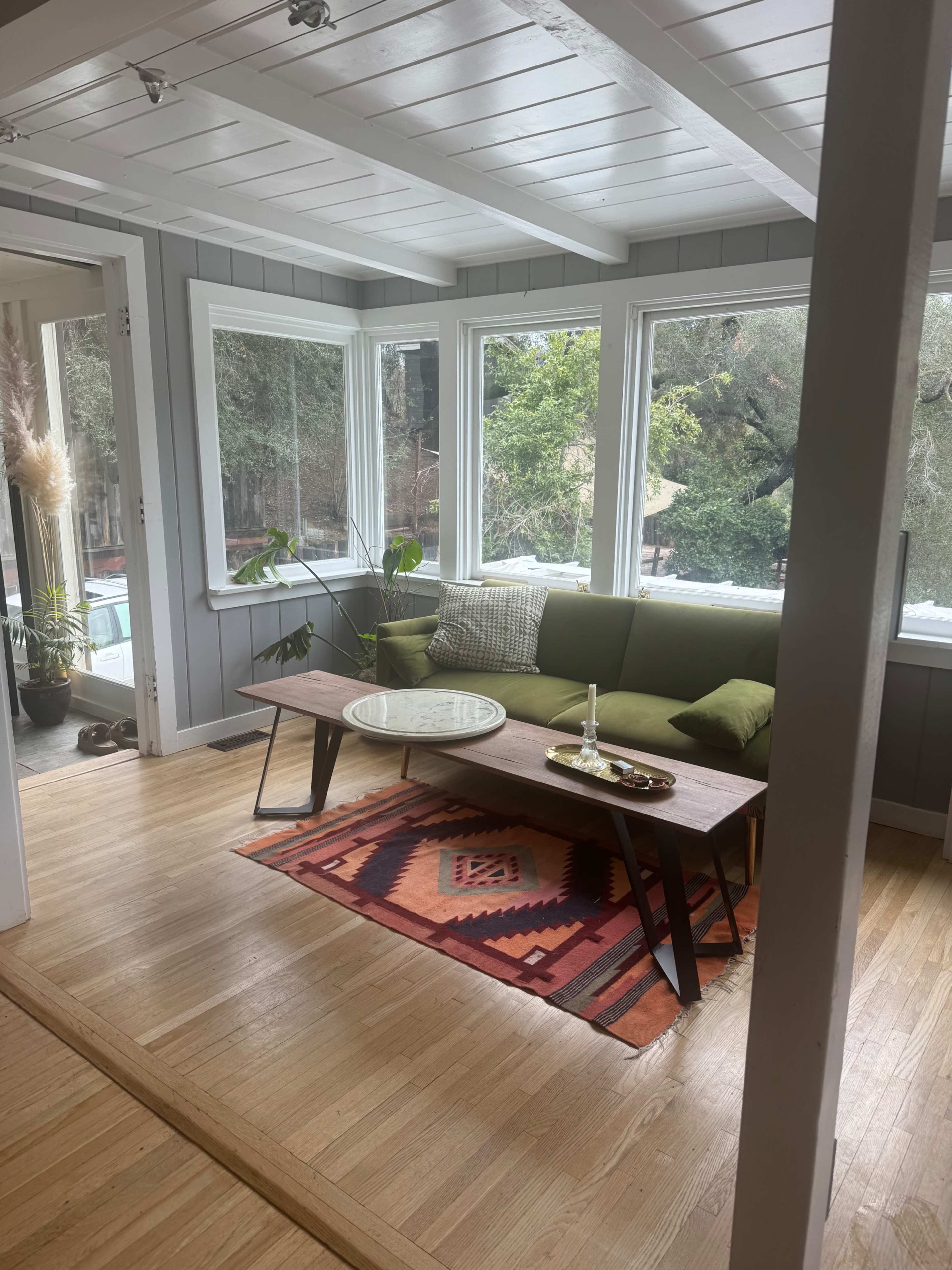 The image shows a cozy sunroom with large windows, a green couch, a wooden coffee table, and a patterned rug on a hardwood floor.