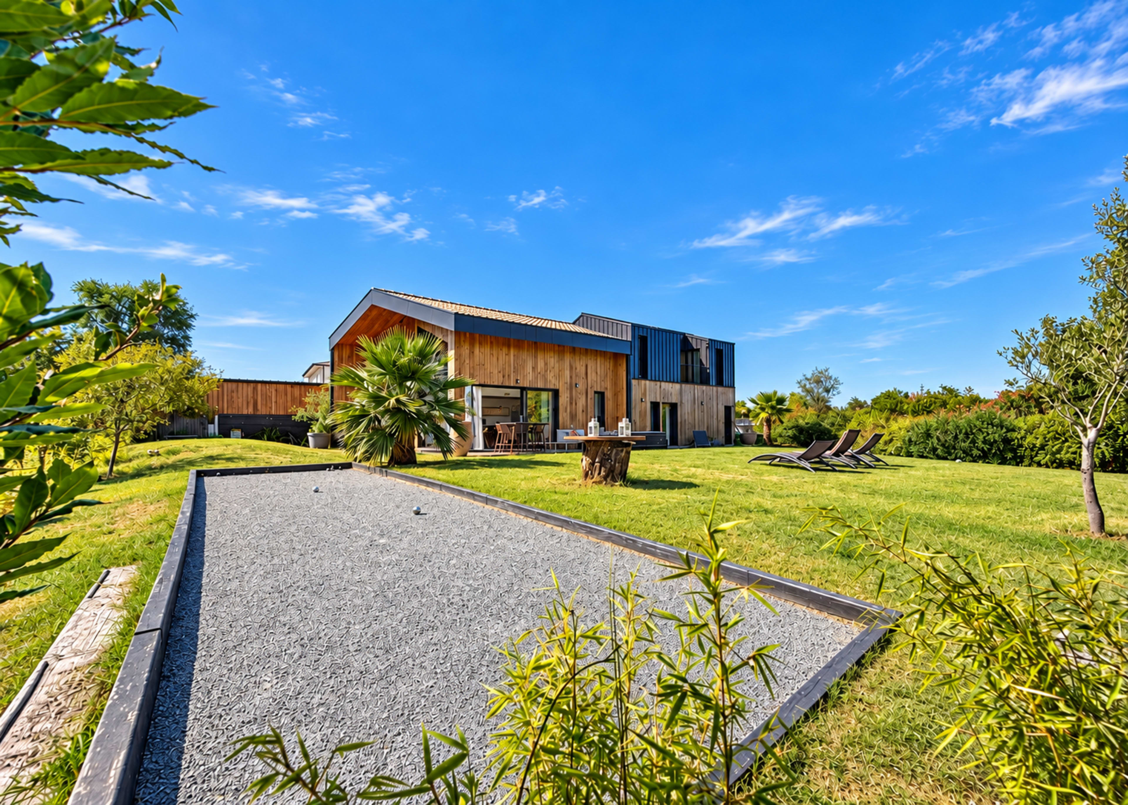 A modern wooden house with a sloped roof sits amidst a lush green yard featuring outdoor seating and a pétanque court.