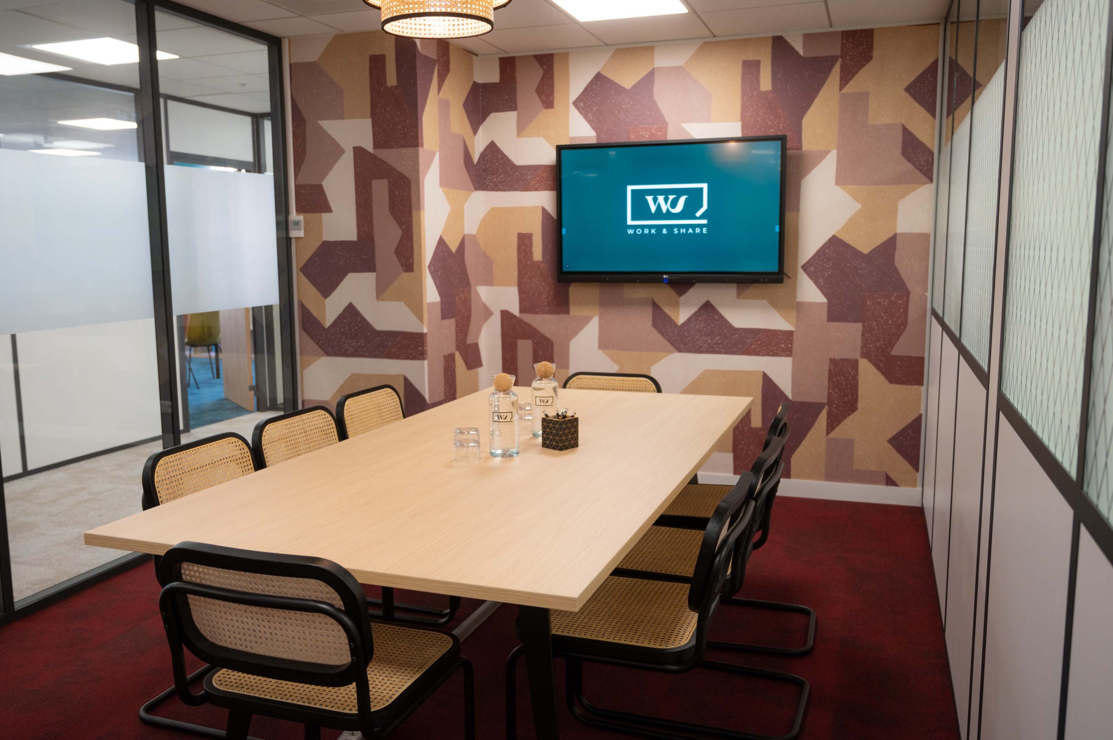 A modern conference room features a wooden table surrounded by six chairs, a large screen displaying the logo "WJ," and a patterned wall in muted colors.