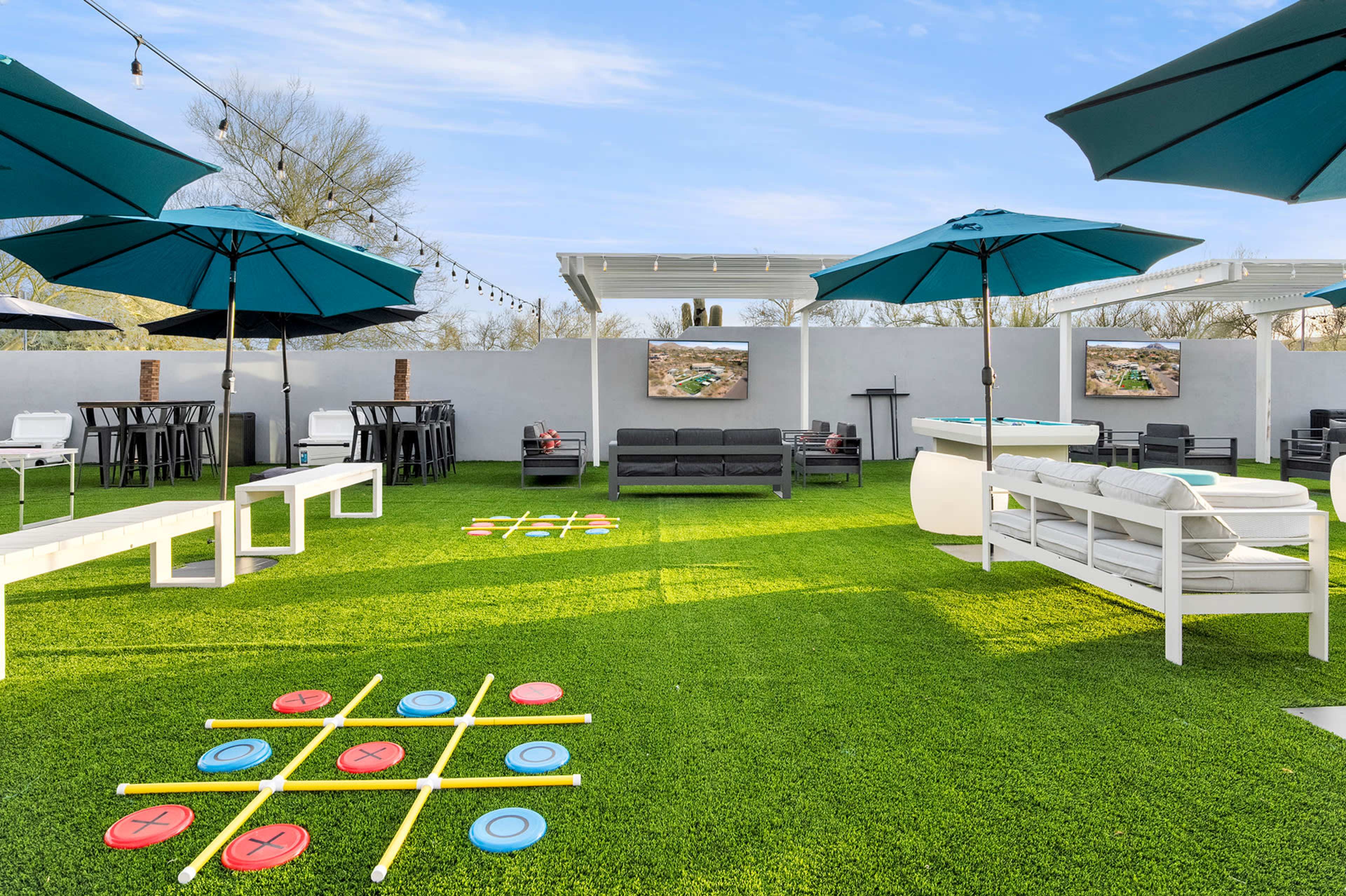 The image shows a spacious outdoor area with green artificial grass, several seating arrangements, large umbrellas, and a game of tic-tac-toe set up on the ground.