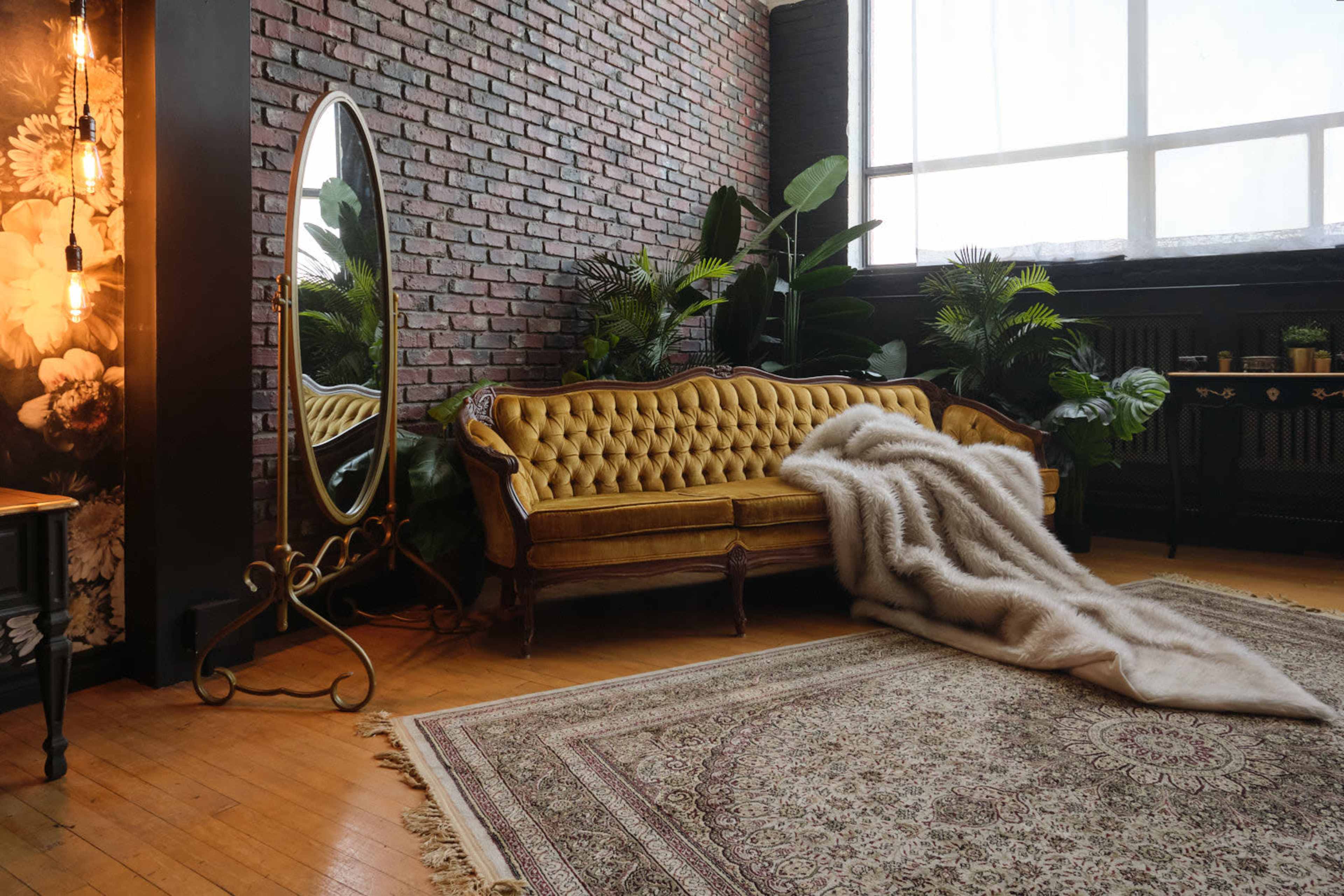 A vintage yellow velvet sofa is positioned next to a large mirror and surrounded by indoor plants in a room with exposed brick walls and a patterned rug.