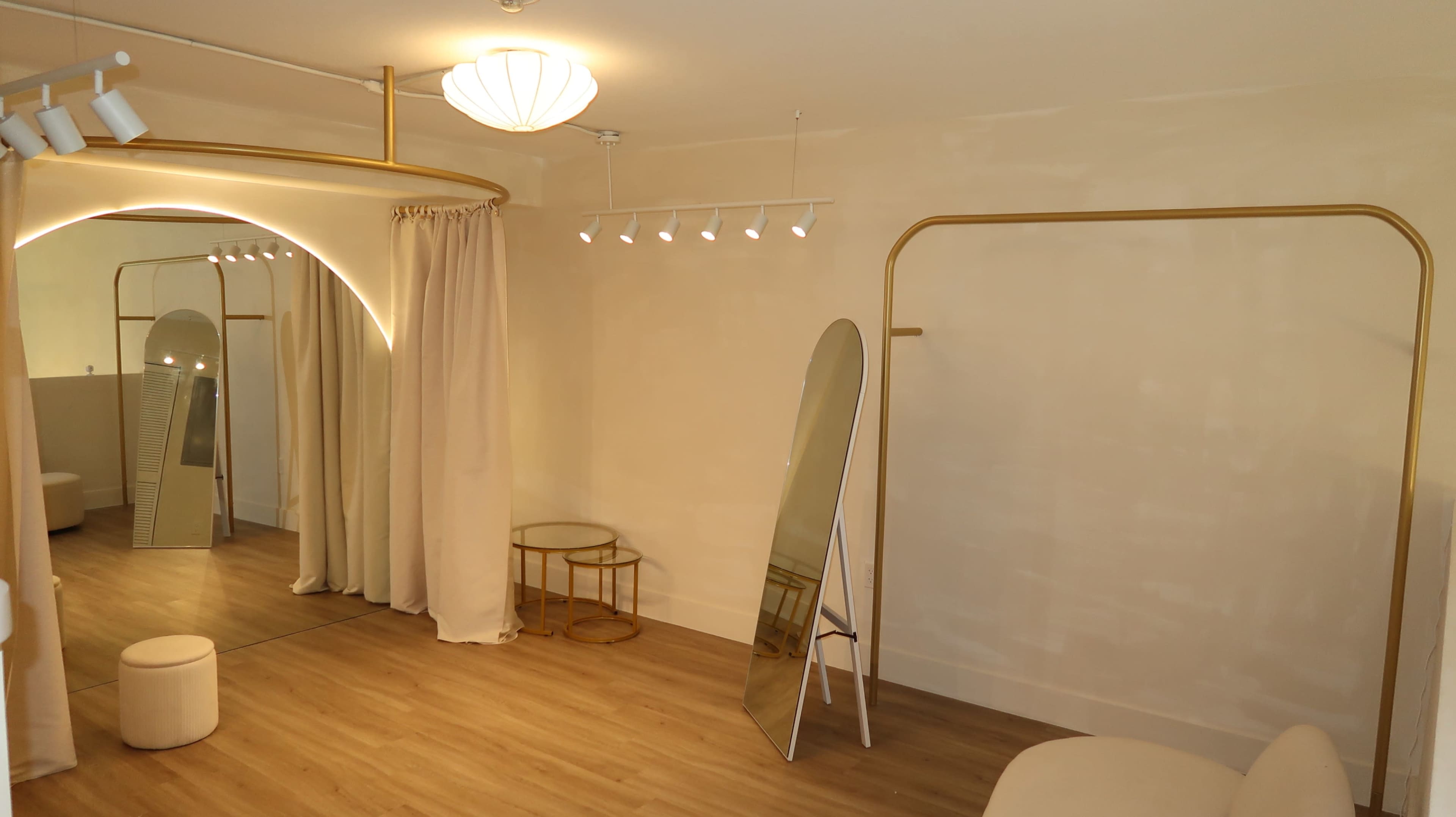 The image displays a minimalistic dressing room with a large mirror, a round stool, a small circular table, and soft, neutral-colored curtains.