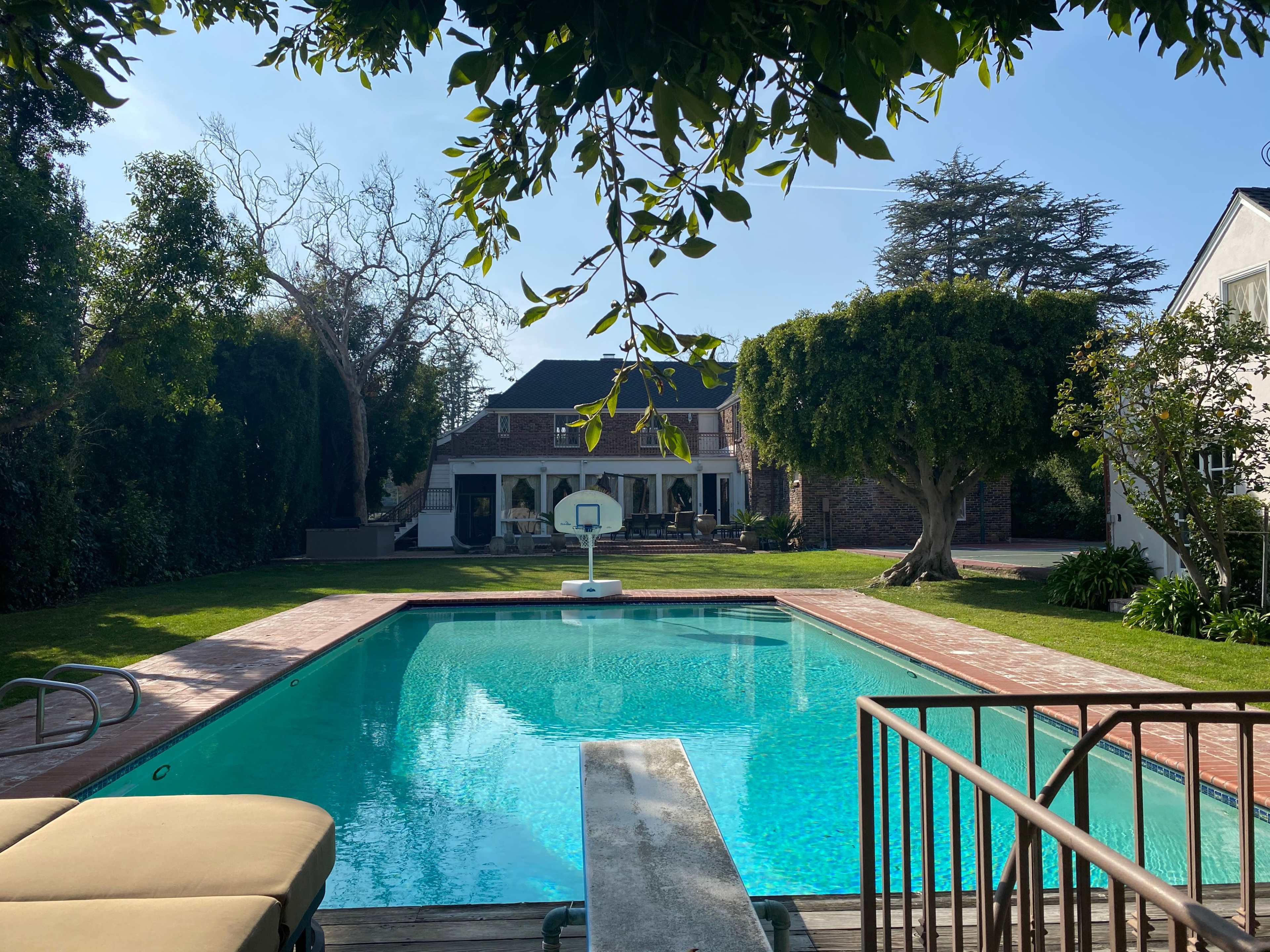 A clear blue swimming pool is framed by lush greenery and a two-story house in the background.