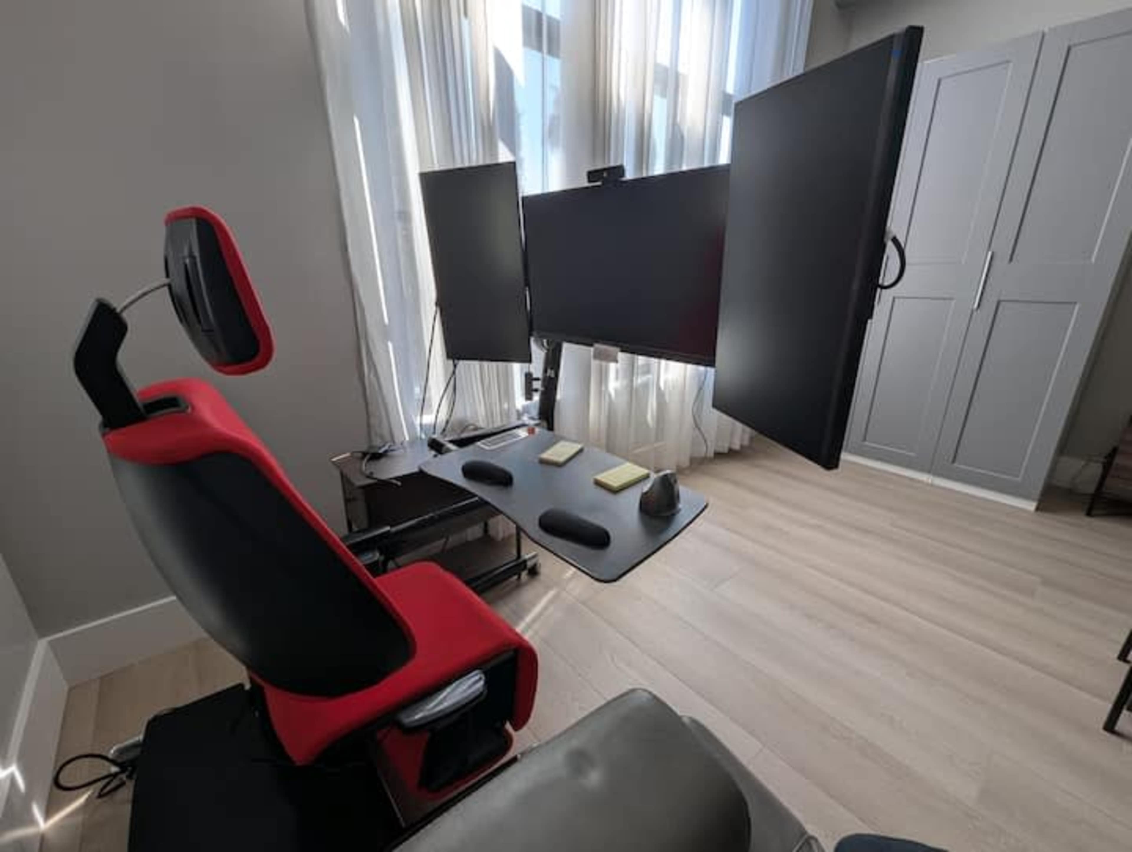 The image shows a modern workspace featuring a red and black gaming chair, two mounted monitors, and a minimalistic desk with sticky notes, all set against a backdrop of large windows with natural light streaming in.