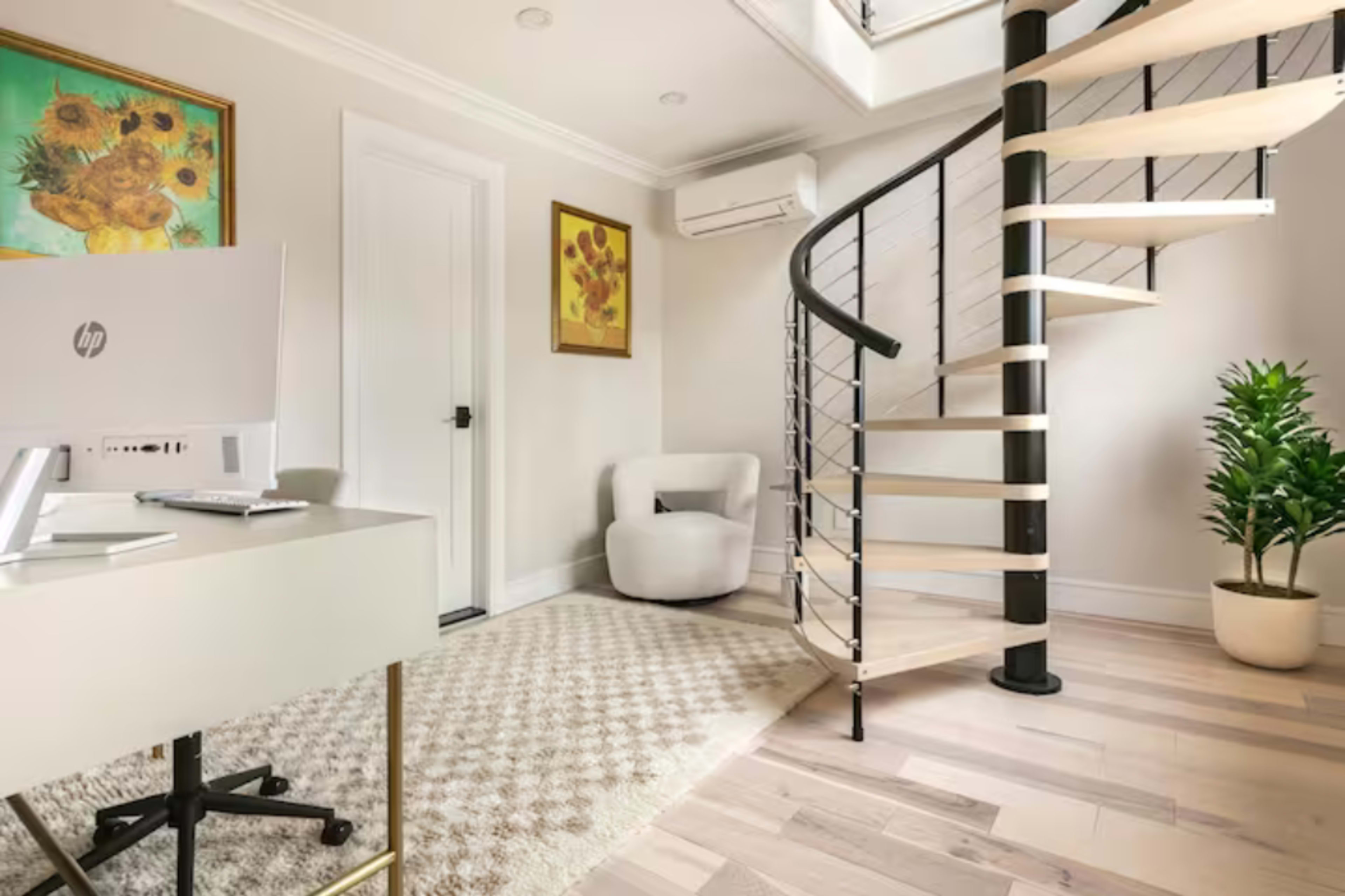 A bright room features a spiral staircase, a small seating area, a desk with a computer, and walls adorned with floral artwork.