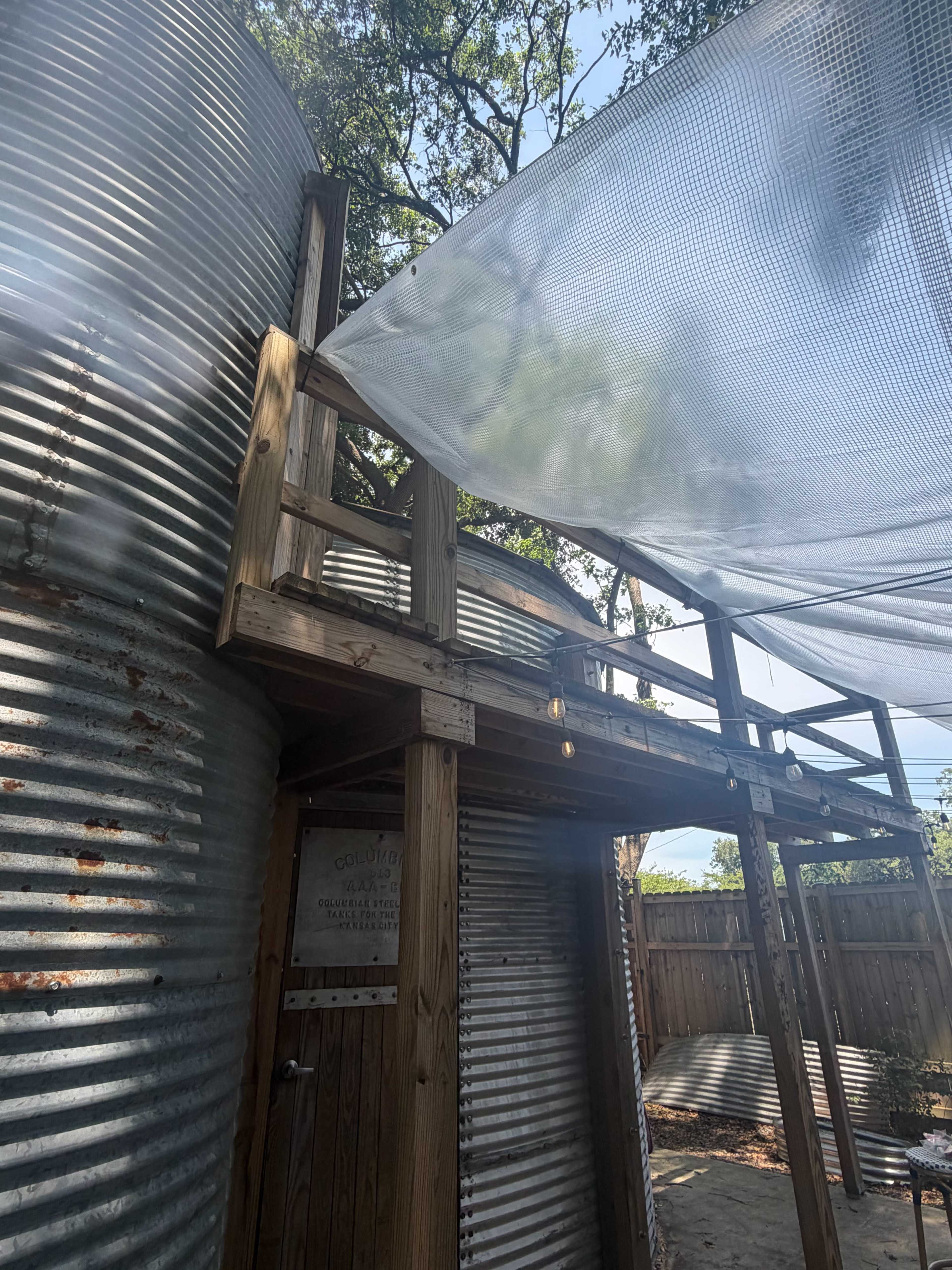 The image shows a wooden deck attached to a corrugated metal structure, surrounded by trees and a shaded area created by a mesh covering.