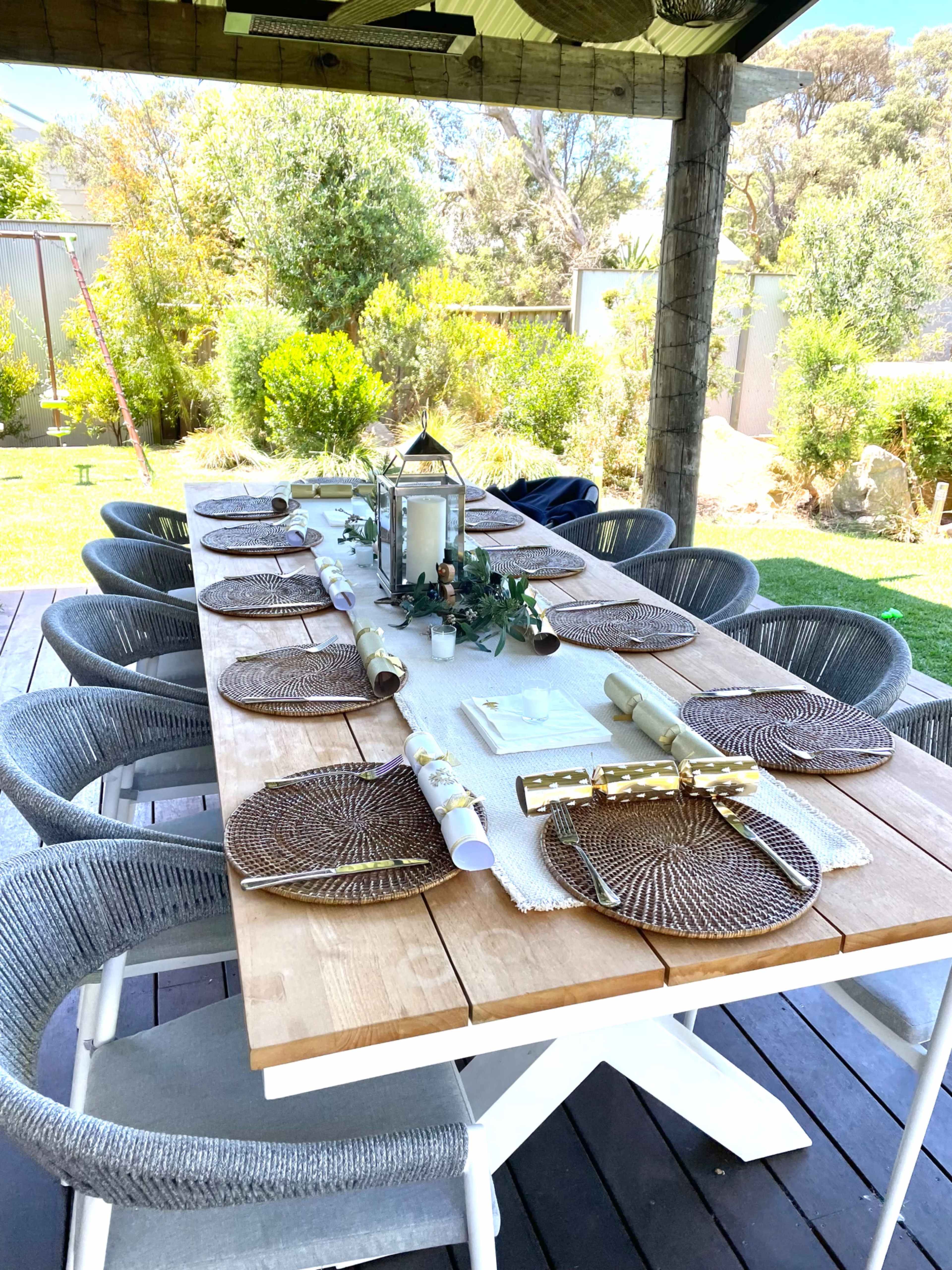 A long wooden dining table is set outdoors with round placemats and decorative items under a shaded area surrounded by greenery.