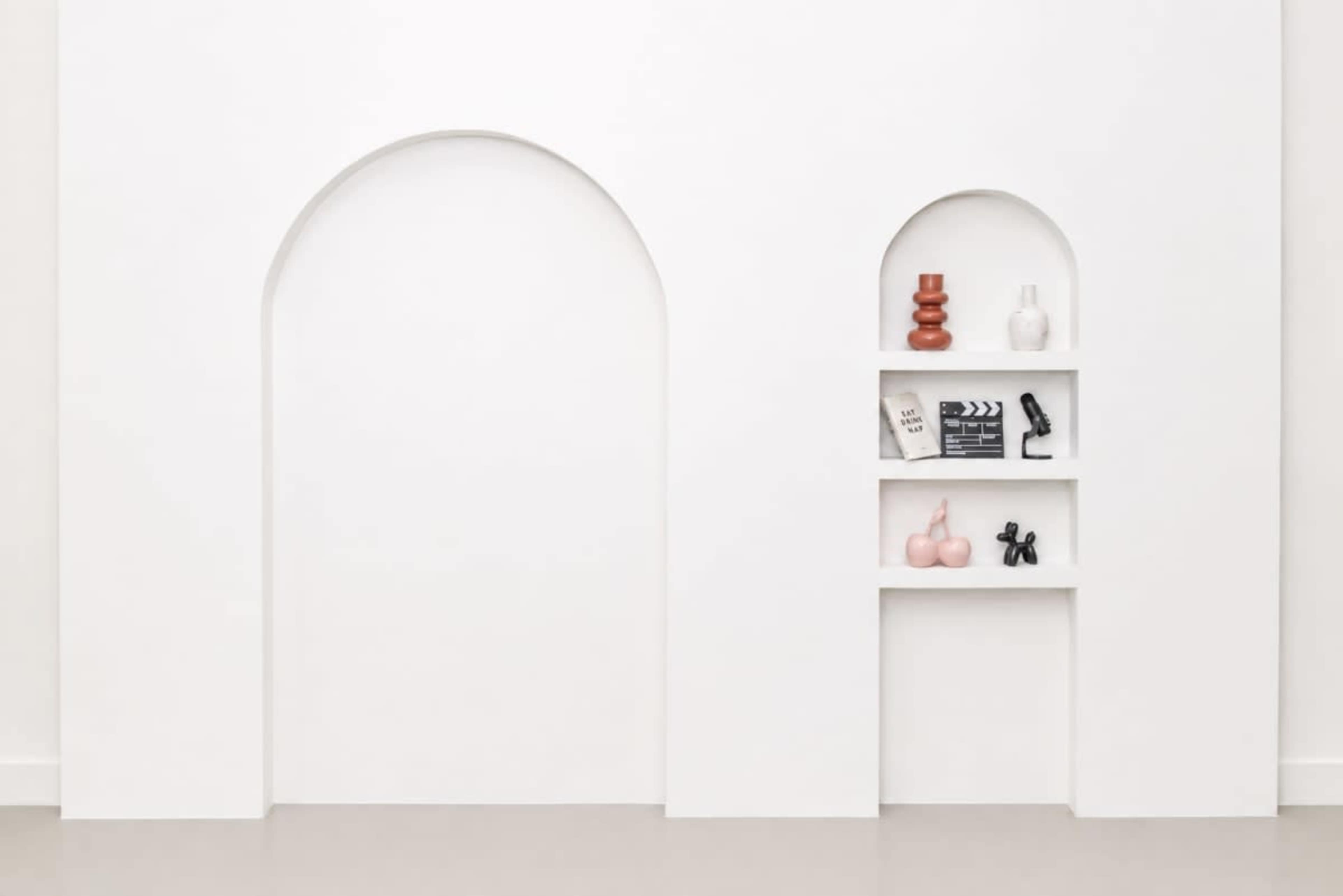 The image features a minimalist white wall with two arch-shaped alcoves, one empty and the other displaying decorative items on a shelf.