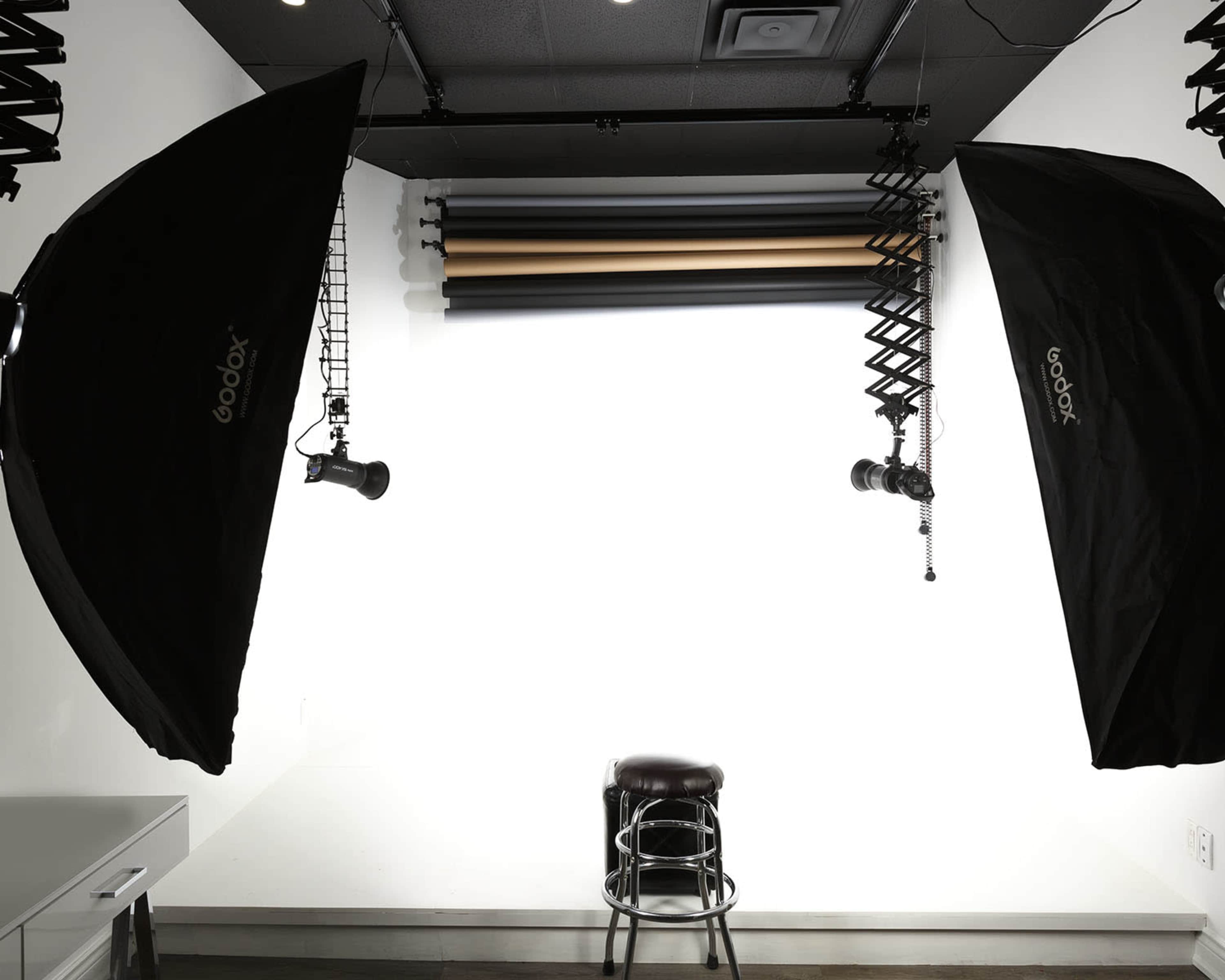 A photography studio setup with two softbox lights, a stool in the center, and a wall mounted with backdrop rolls in various colors.