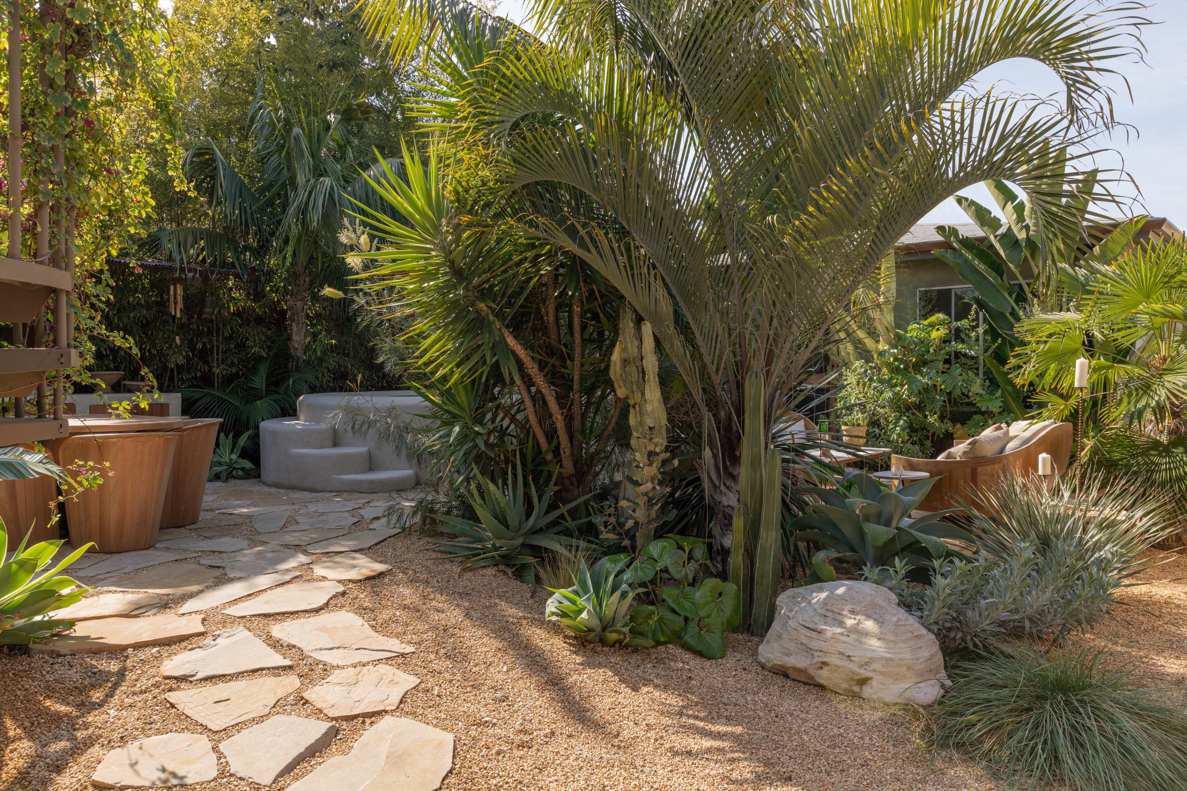 The image shows a landscaped garden featuring stone pathways, various plants, and a small hot tub surrounded by lush greenery.