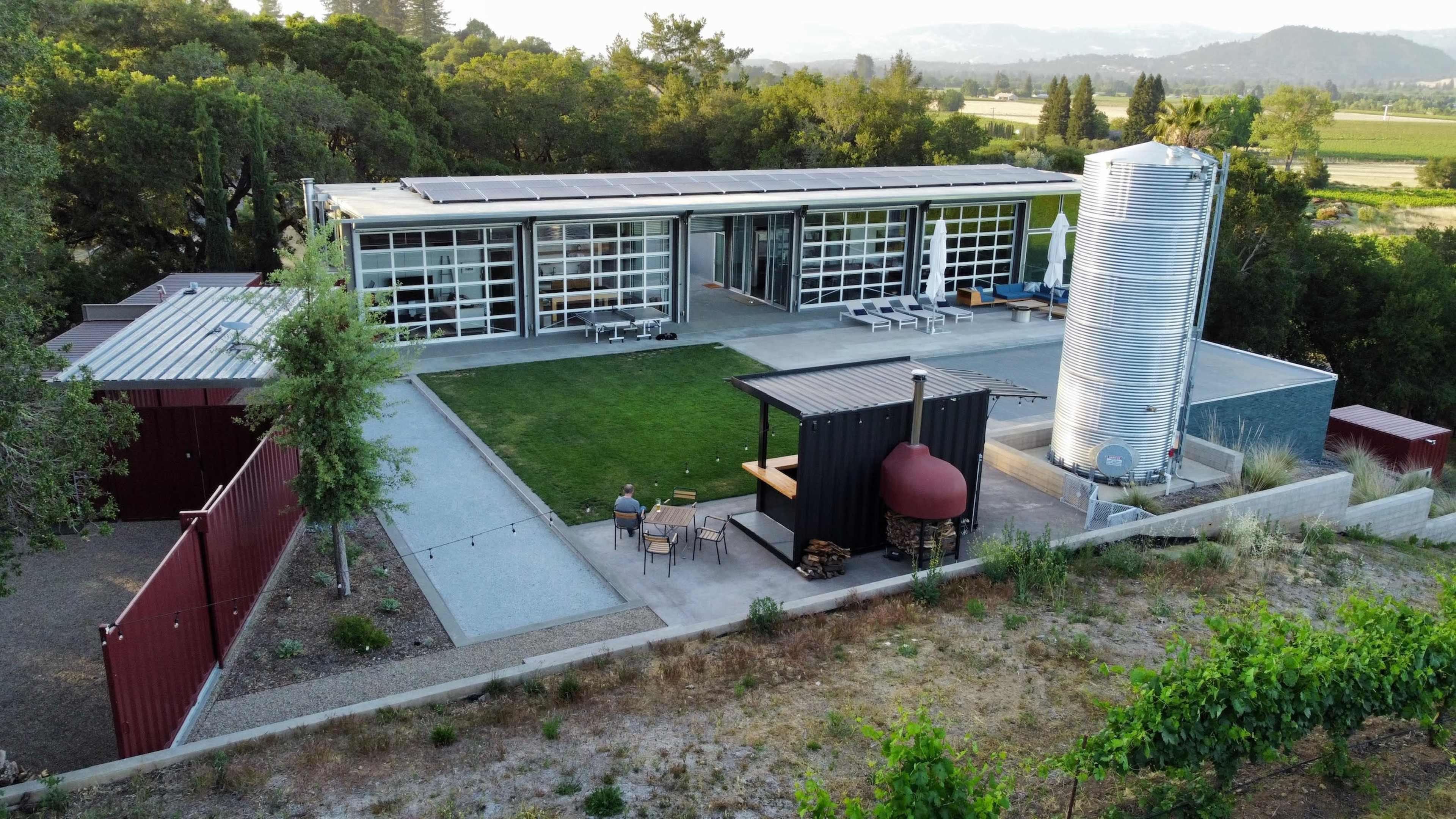 A modern building with large glass windows and solar panels is surrounded by a landscaped area, featuring a seating space and a nearby metal water tank.