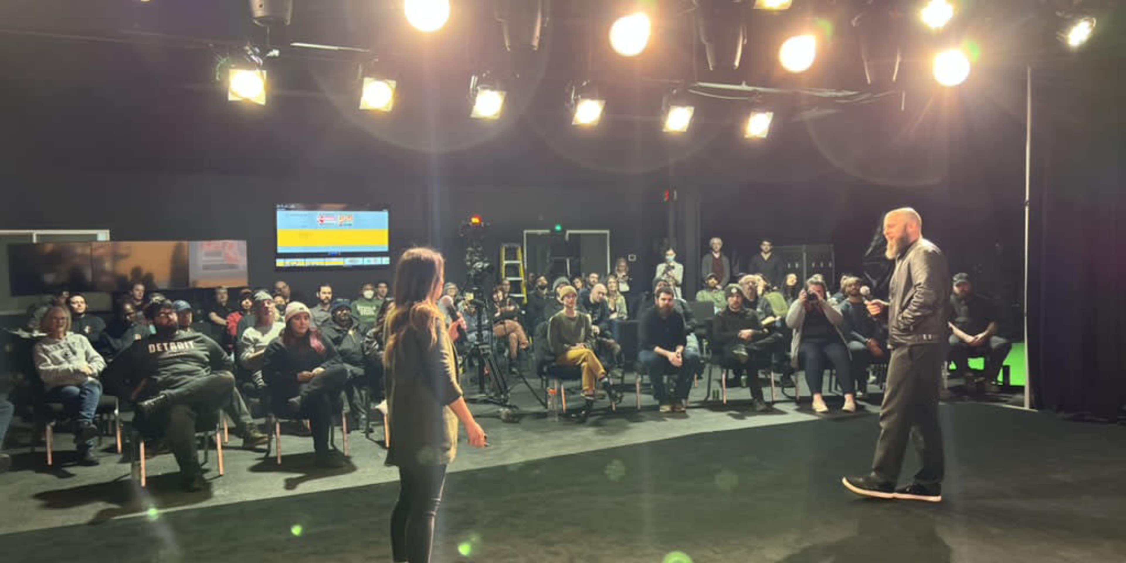 A speaker addresses an audience in a brightly lit studio.