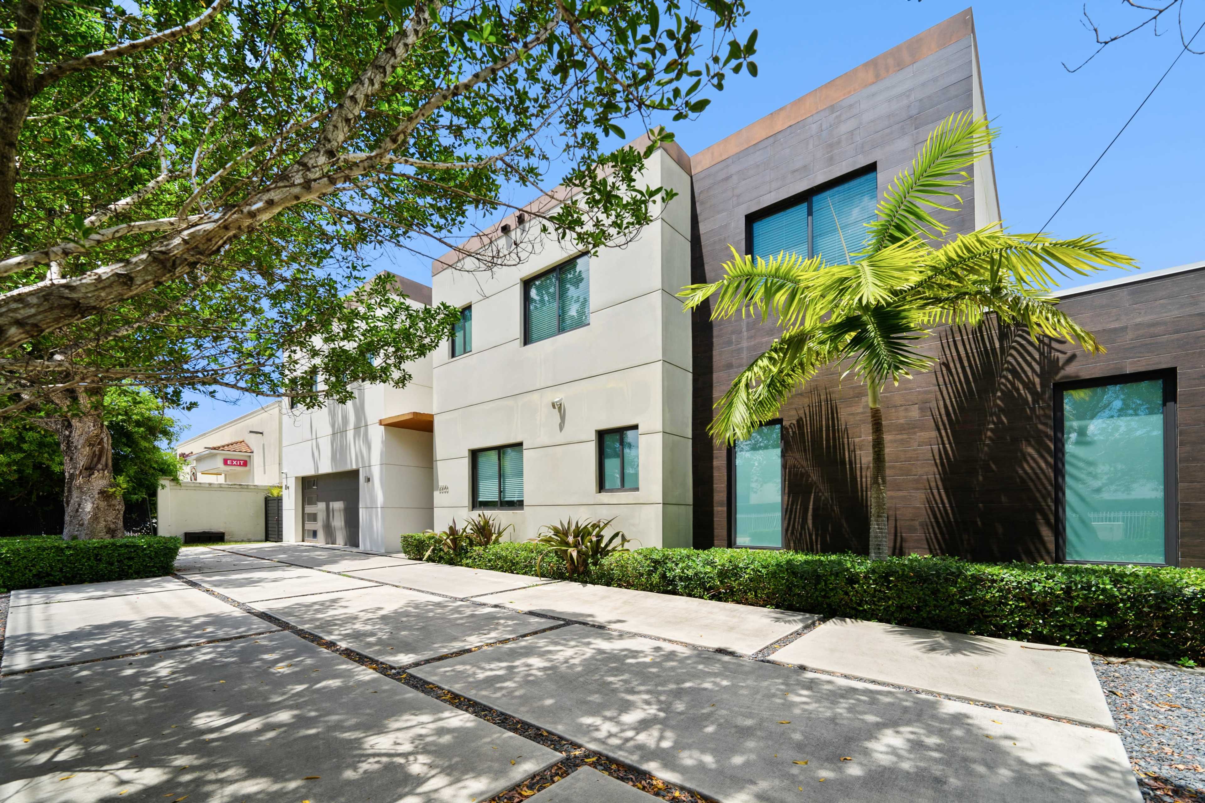 A modern two-story house with large windows, a landscaped front yard, and a concrete driveway lined with greenery.