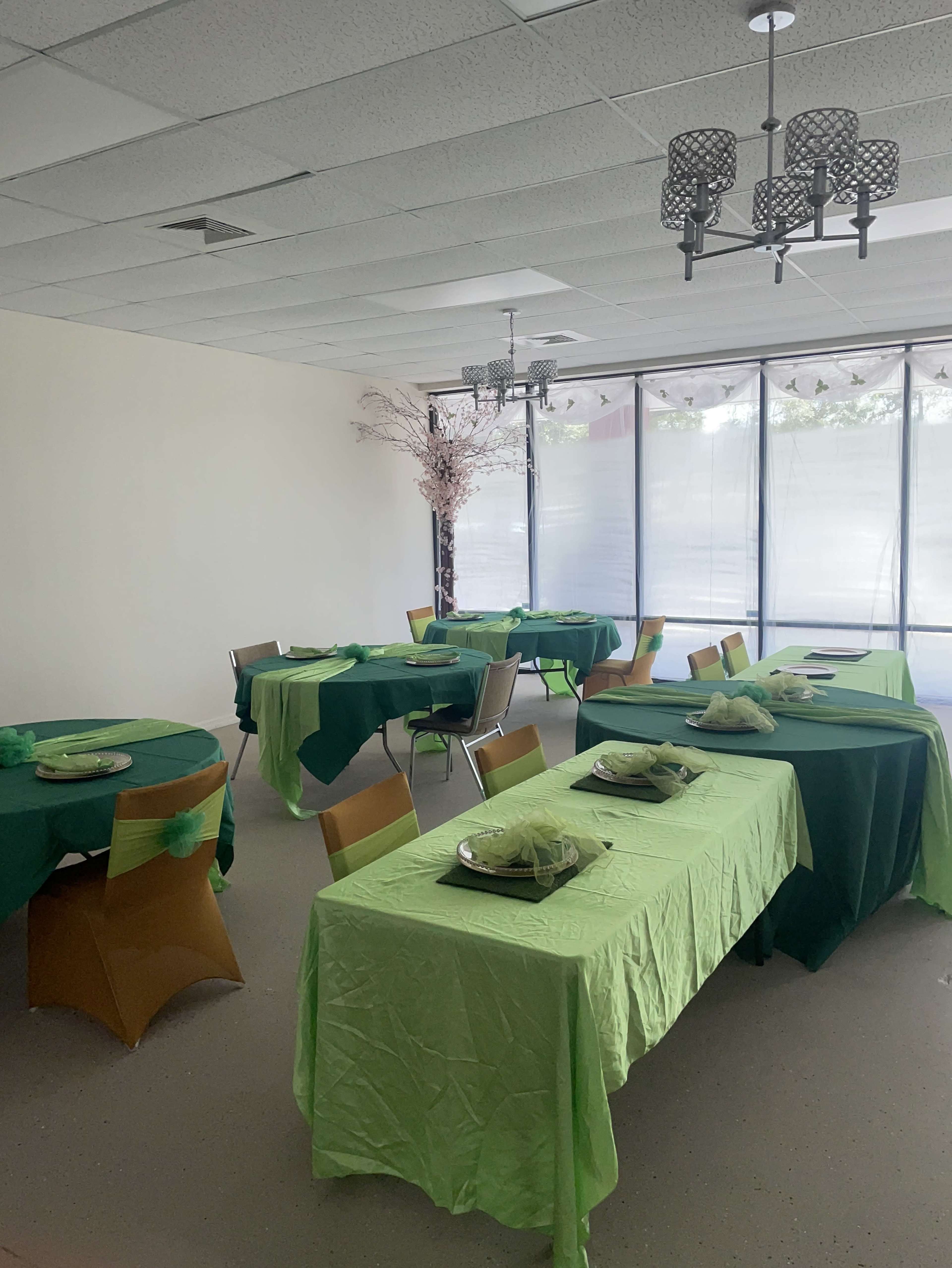 A spacious room set up for an event with several tables covered in green and light green tablecloths, each arranged with plates and decorative items.