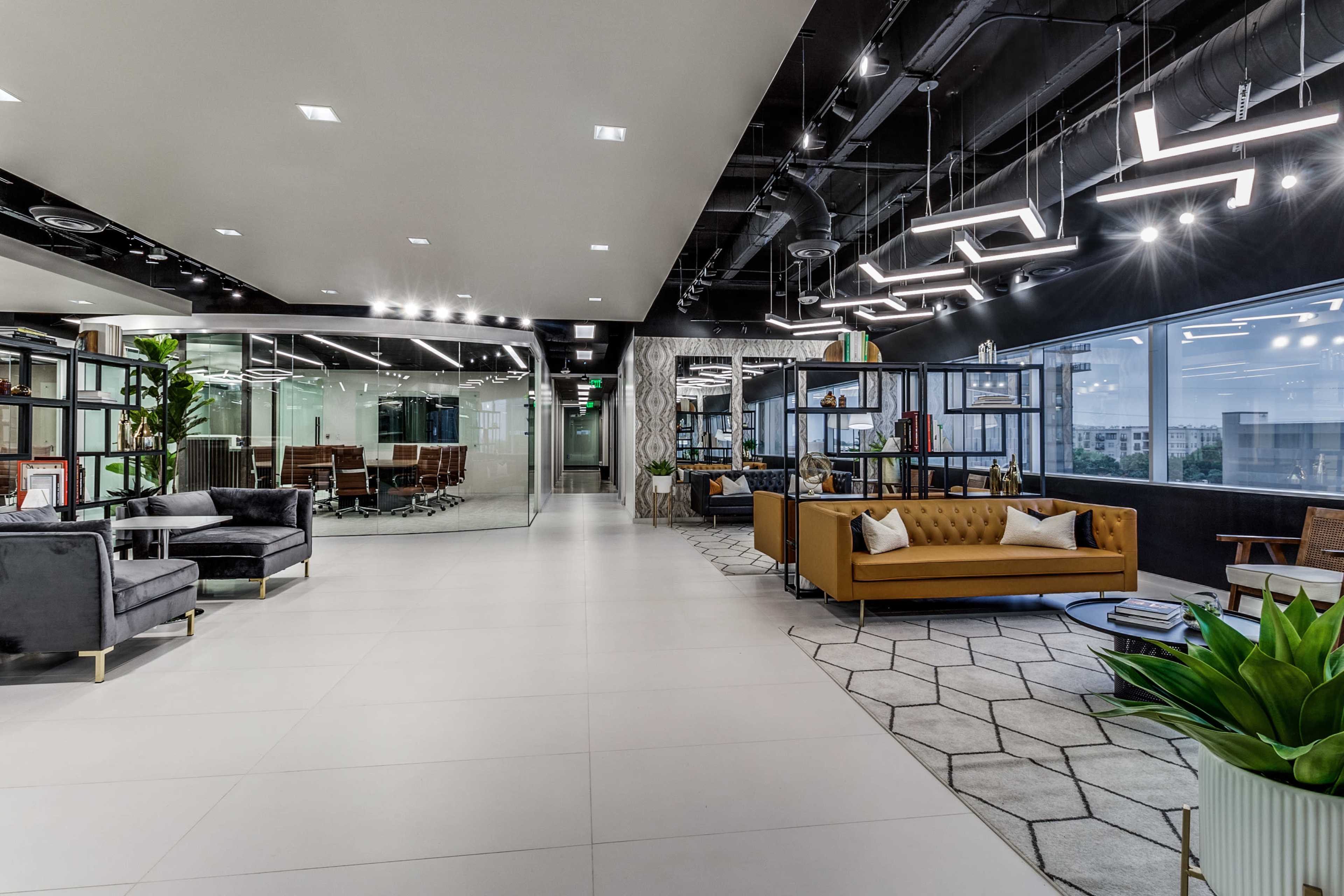 A modern office interior featuring a spacious seating area with a brown sofa and contemporary lighting fixtures, adjacent to glass-walled meeting rooms.