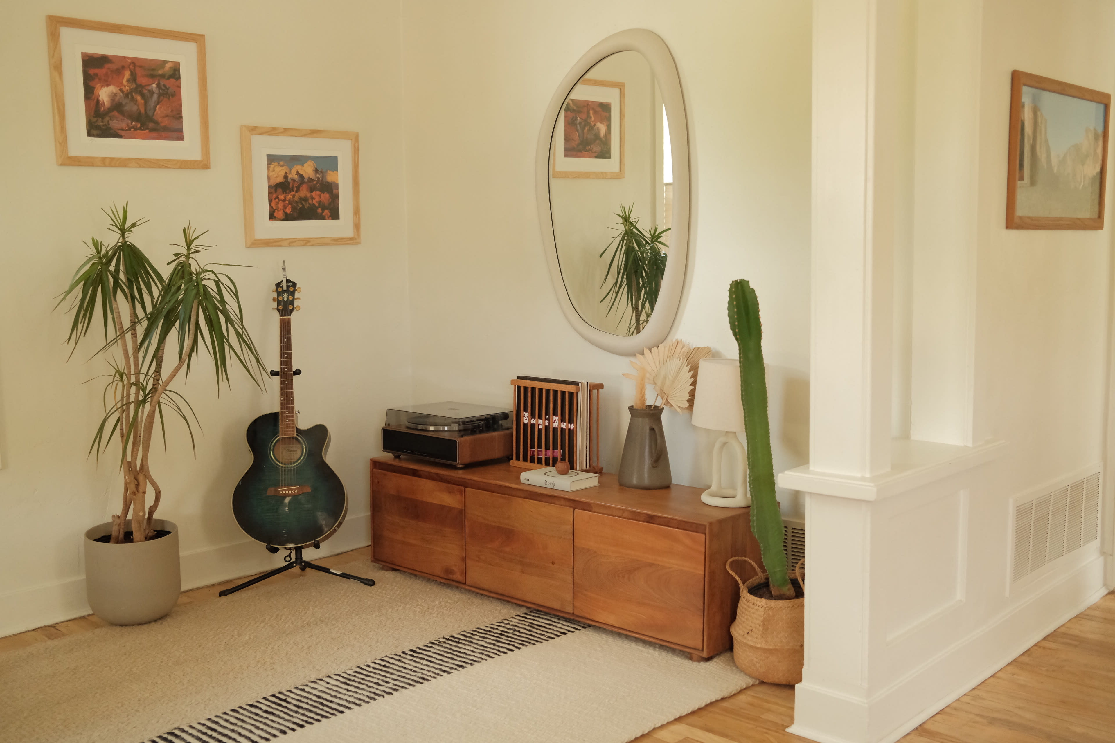 The image shows a cozy corner of a room featuring a wooden console table, a guitar, plants, framed artwork on the walls, and decorative items.