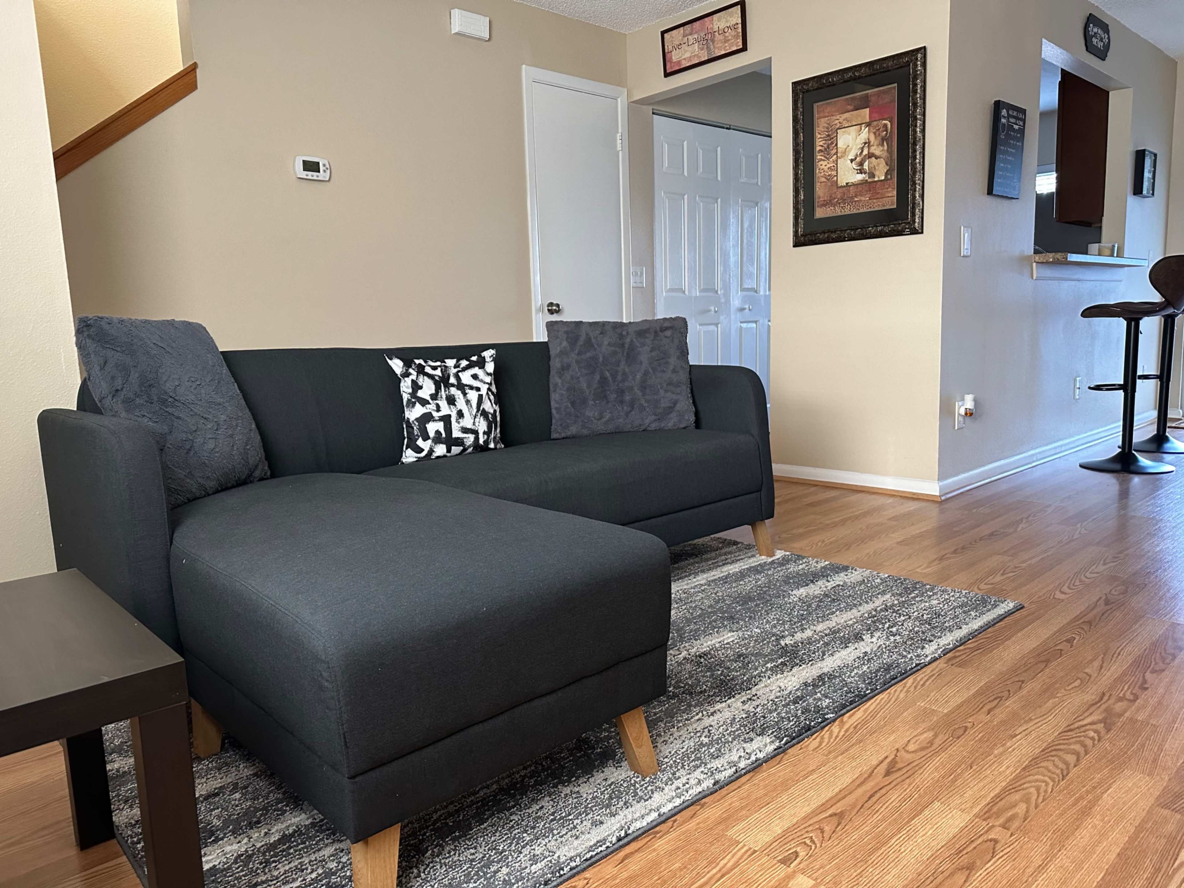 The image shows a modern living room featuring a black sectional sofa with decorative pillows, a patterned rug, and a bar area in the background.
