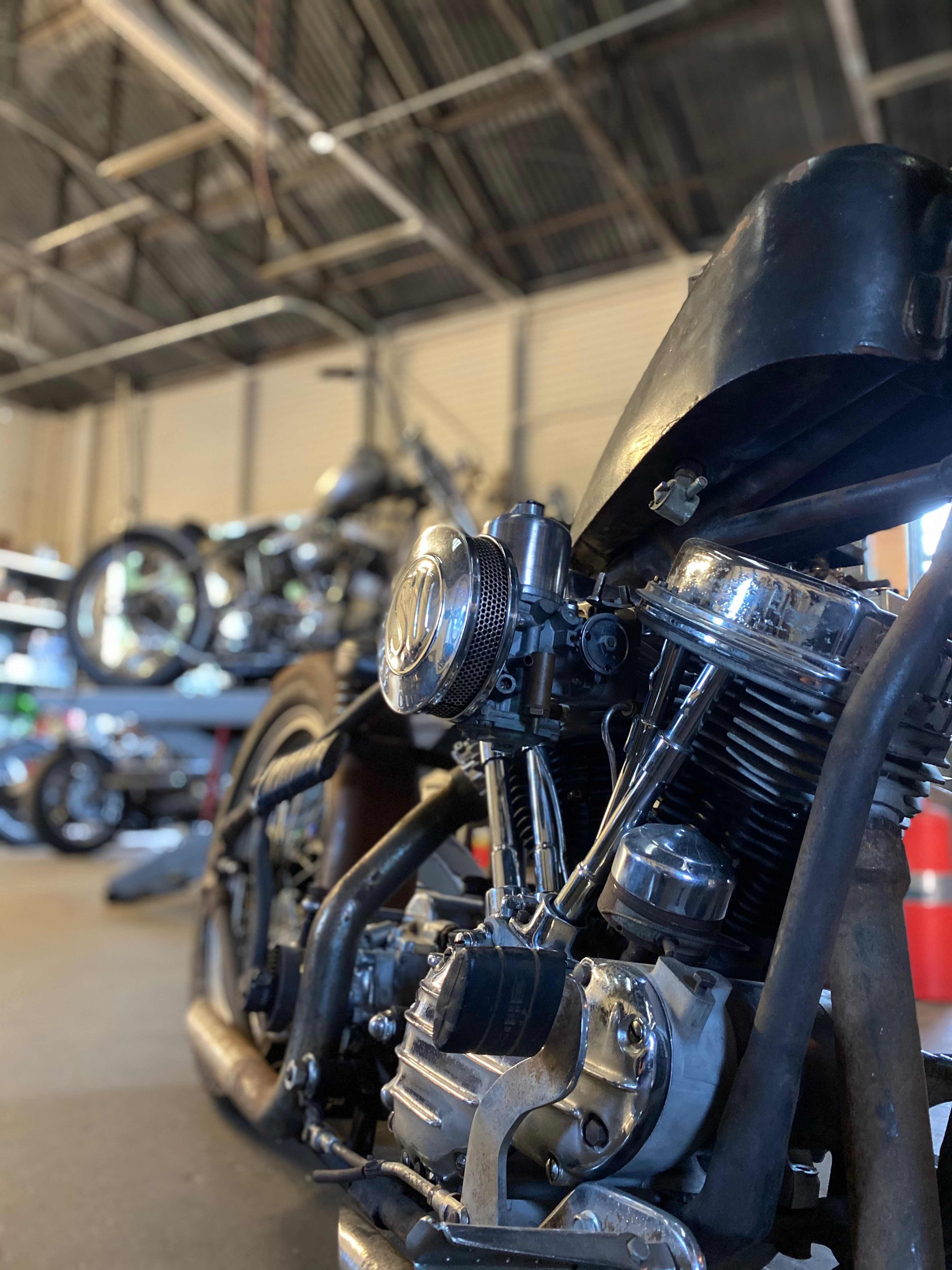 The image shows a close-up view of a vintage motorcycle engine in a garage filled with other motorcycles in the background.