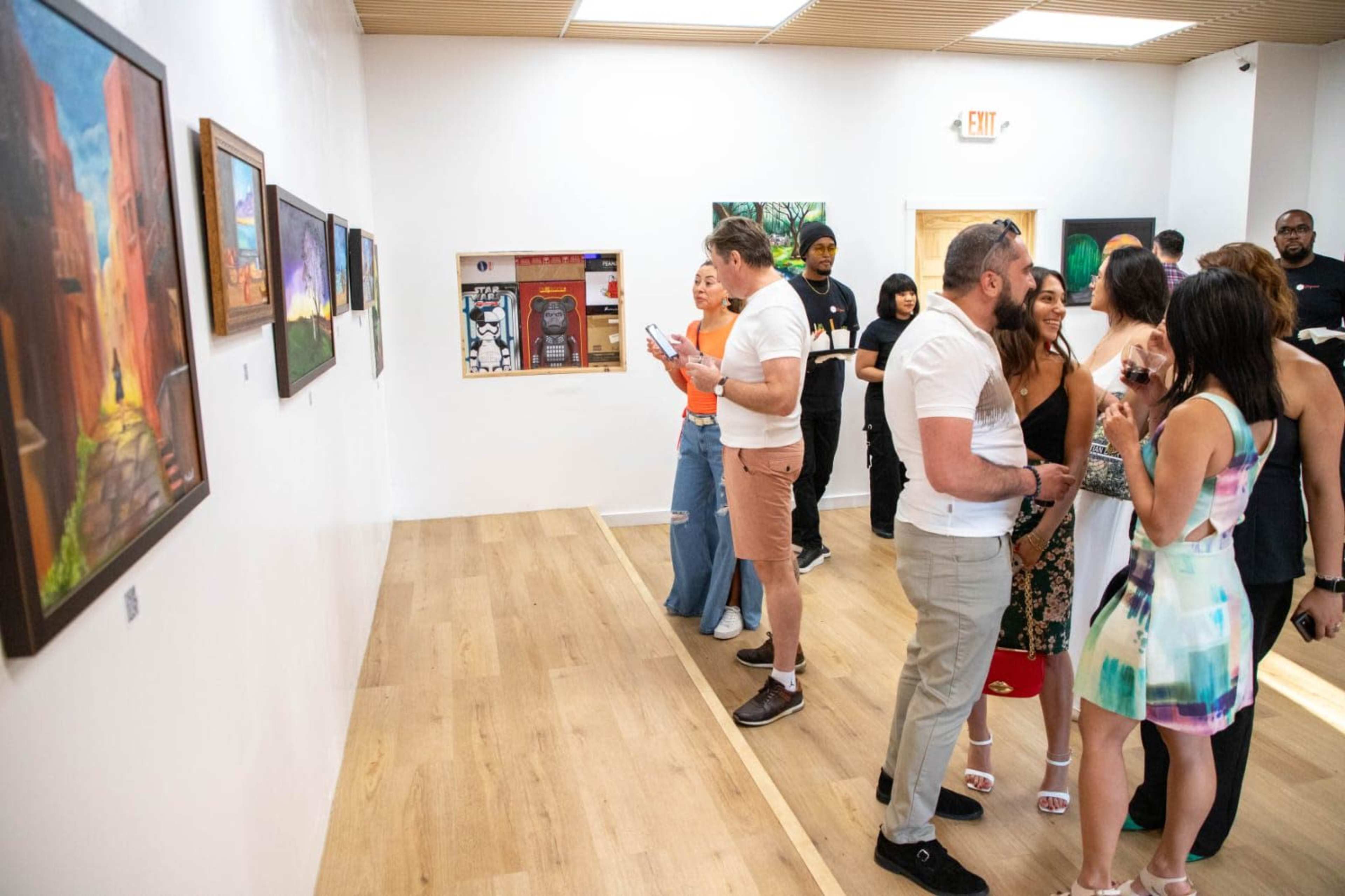 A group of people is gathered in an art gallery, engaging with various paintings displayed on the walls.
