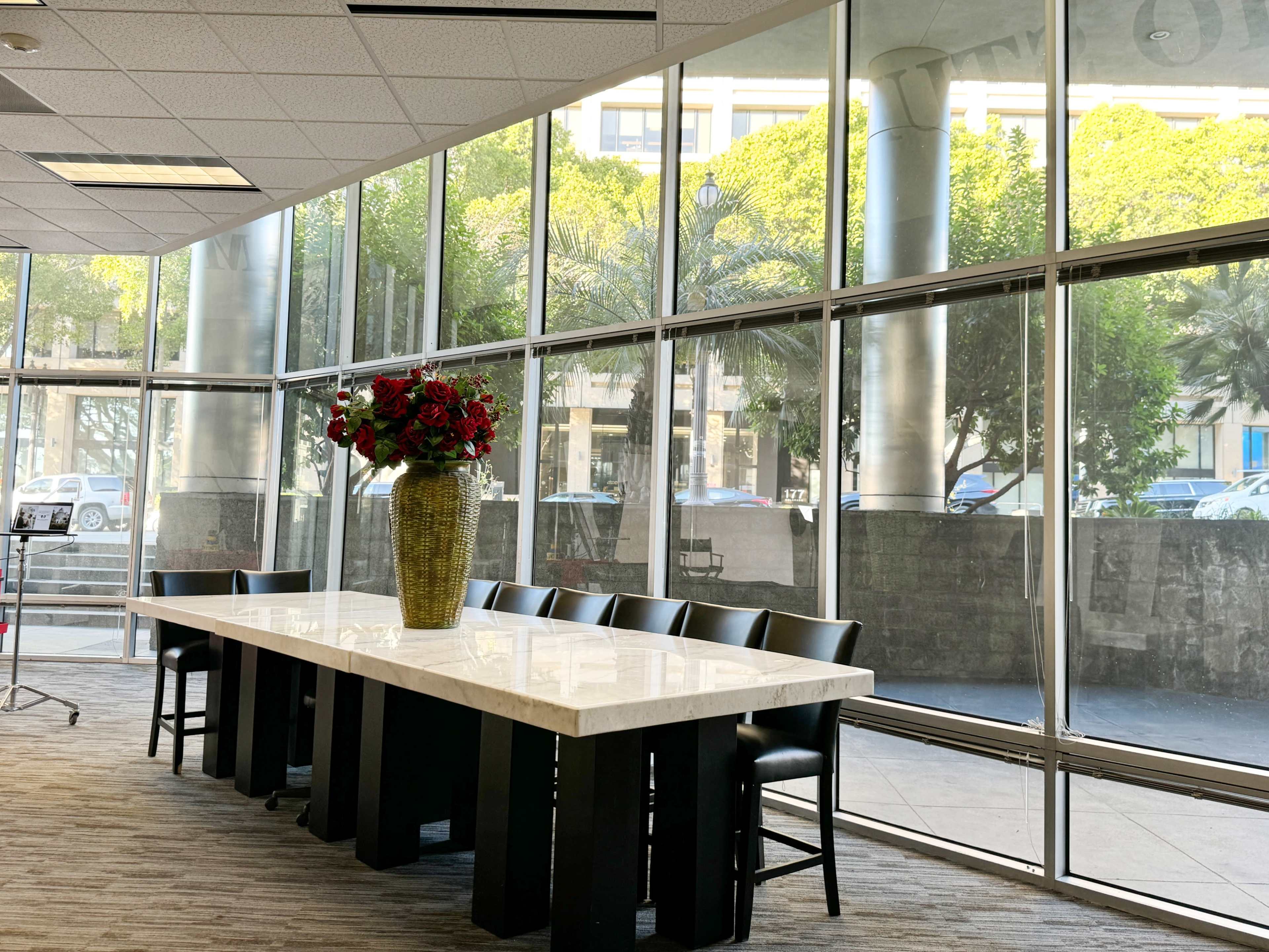 A long white table with black chairs is placed near large glass windows, showcasing greenery and outdoor scenery.