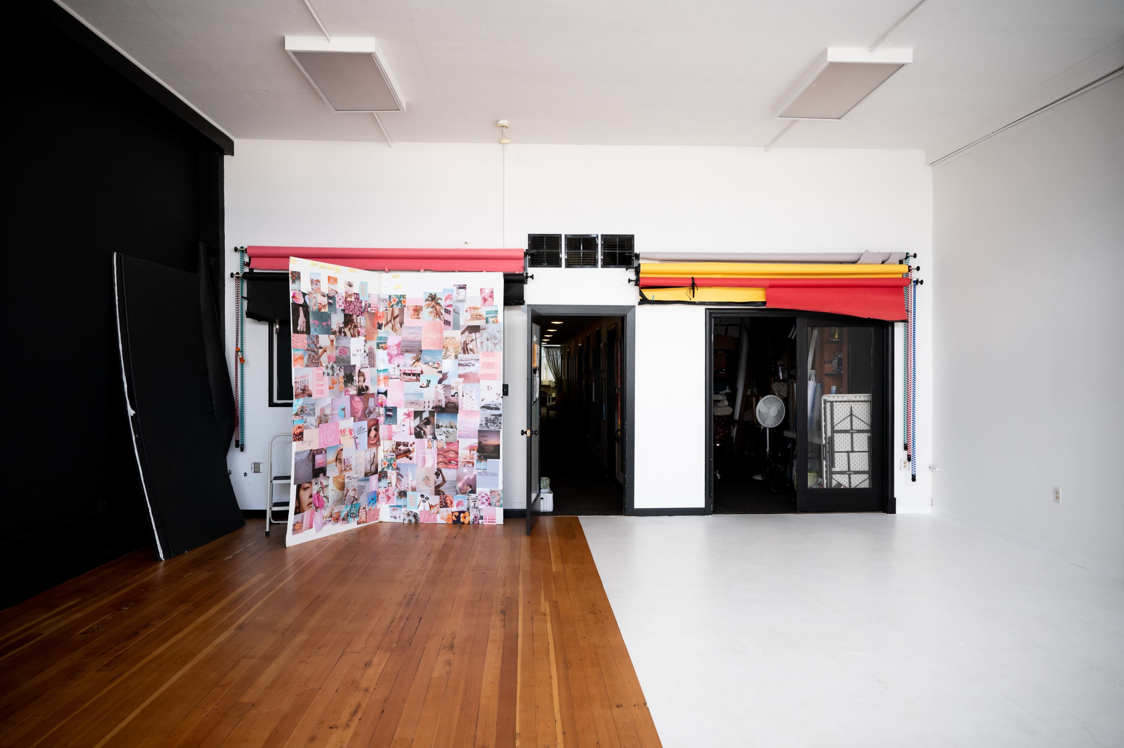 A spacious studio with a wooden floor, featuring a collage panel on the left and a brightly lit entrance to another room adorned with colorful backdrops.