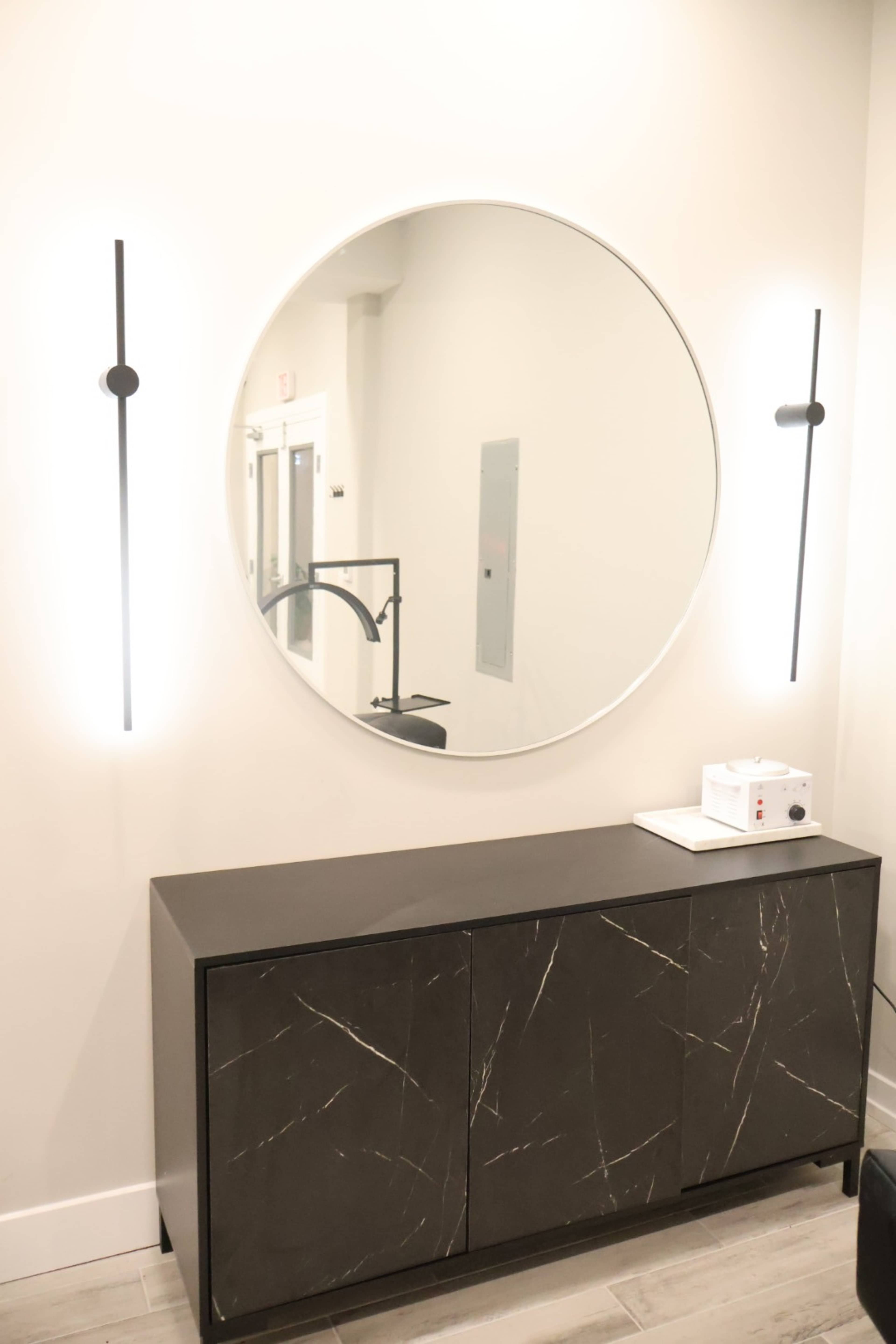 A circular mirror is mounted on a wall above a black cabinet with a marble-like finish, flanked by two sleek wall sconces.