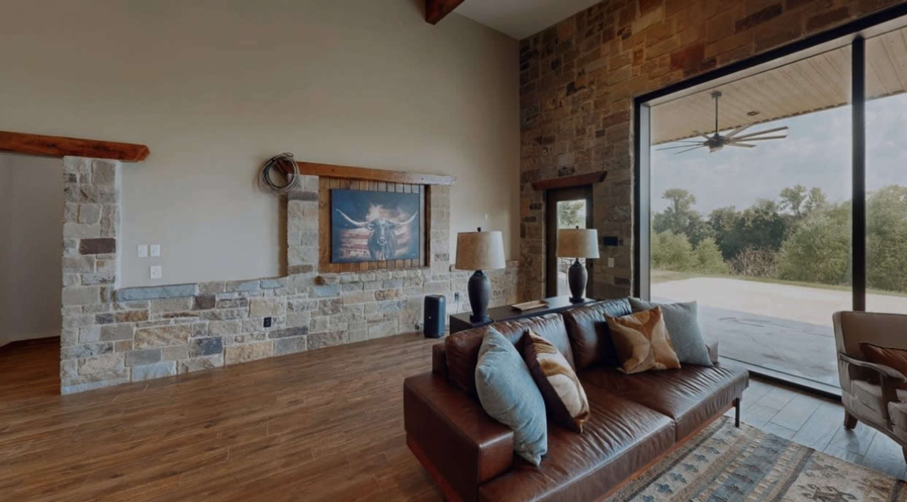The image depicts a modern living room featuring a leather sofa, wooden beams, stone walls, and large windows that open to an outdoor view.