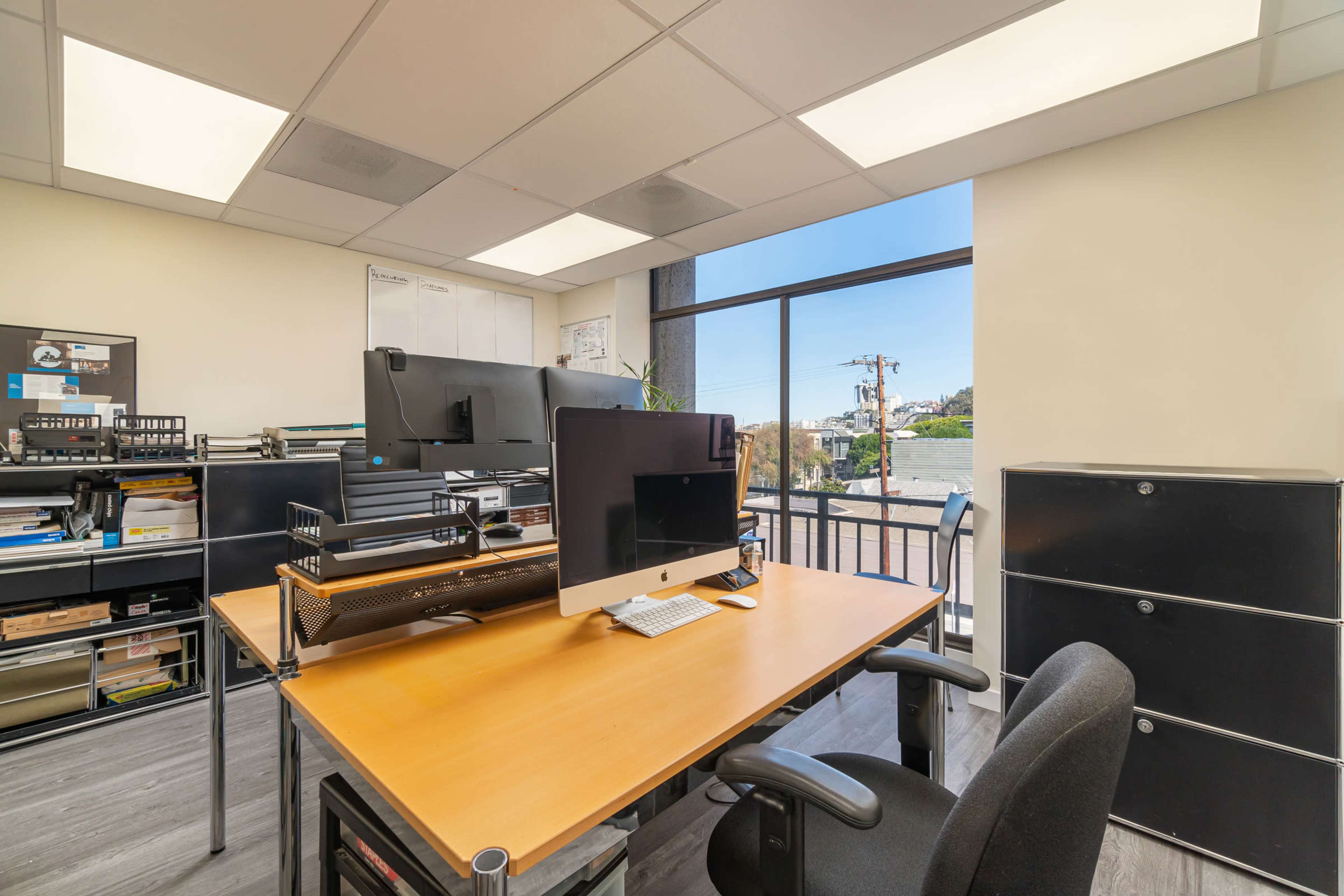 A modern office space with a wooden desk, computer setup, and shelves filled with office supplies, all illuminated by natural light from a large window.