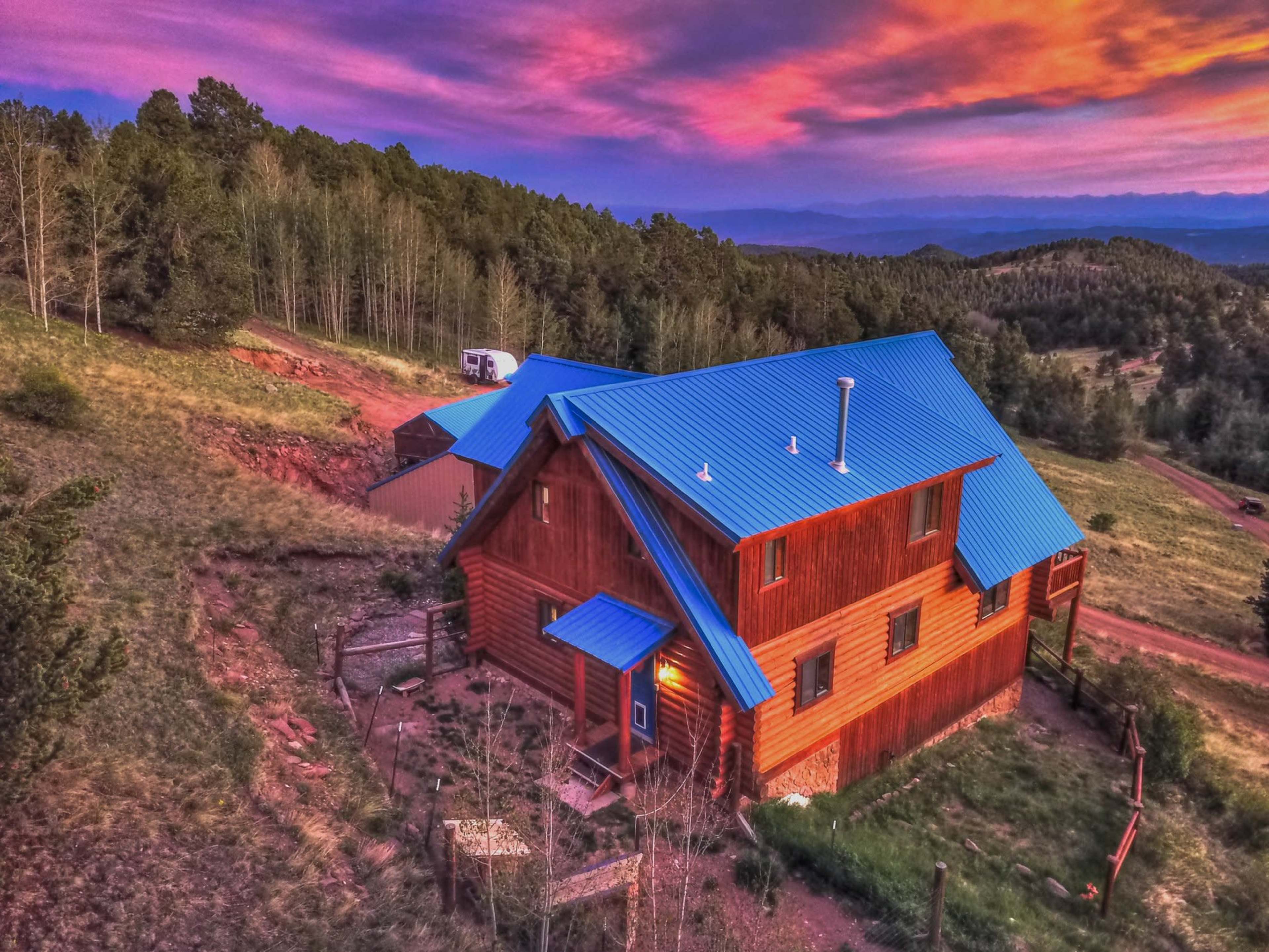 A wooden house with a blue roof is situated on a hillside surrounded by trees under a vibrant sunset sky.