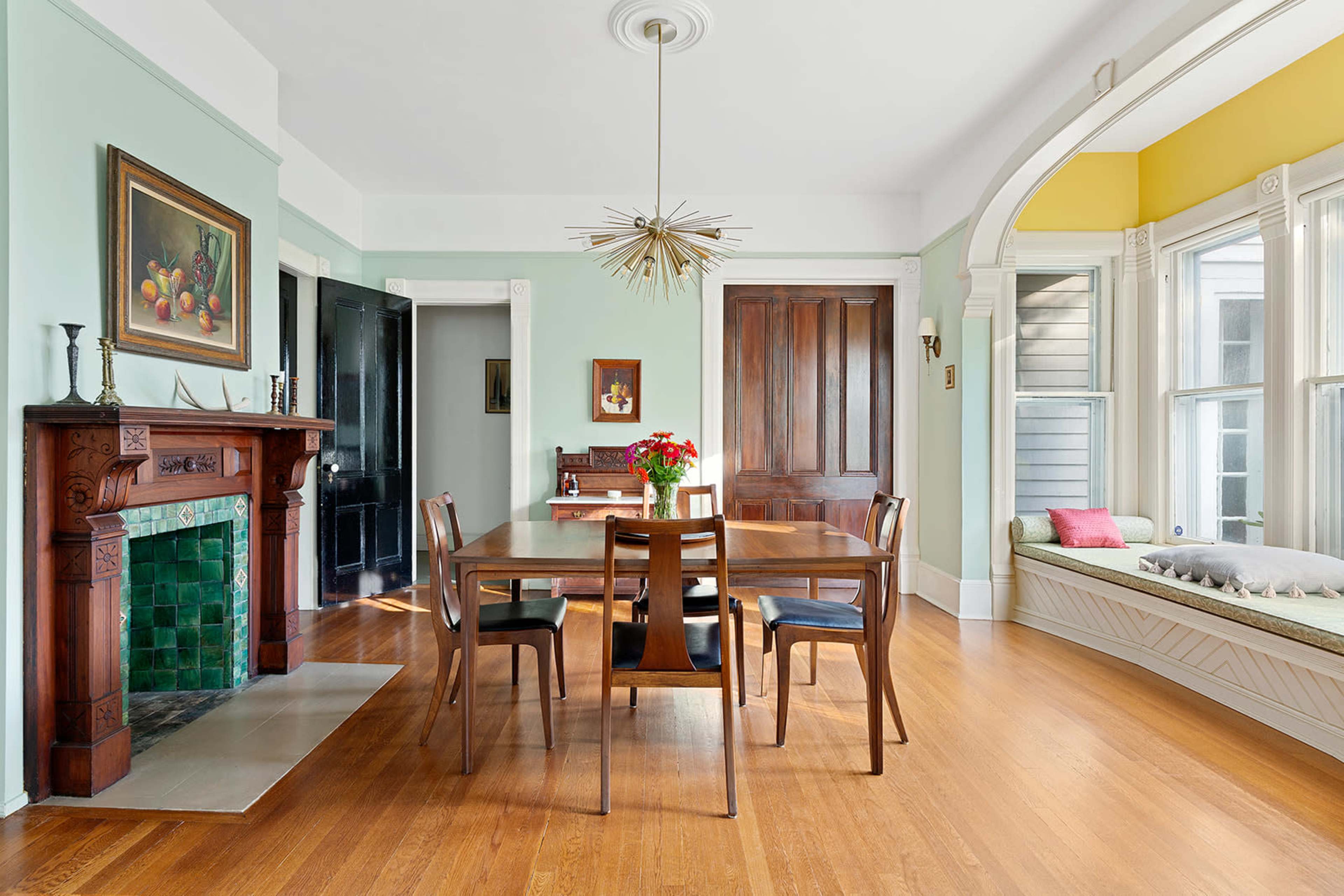 A dining room features a wooden table surrounded by chairs, a fireplace with green tiles, and a large window seat bathed in natural light.
