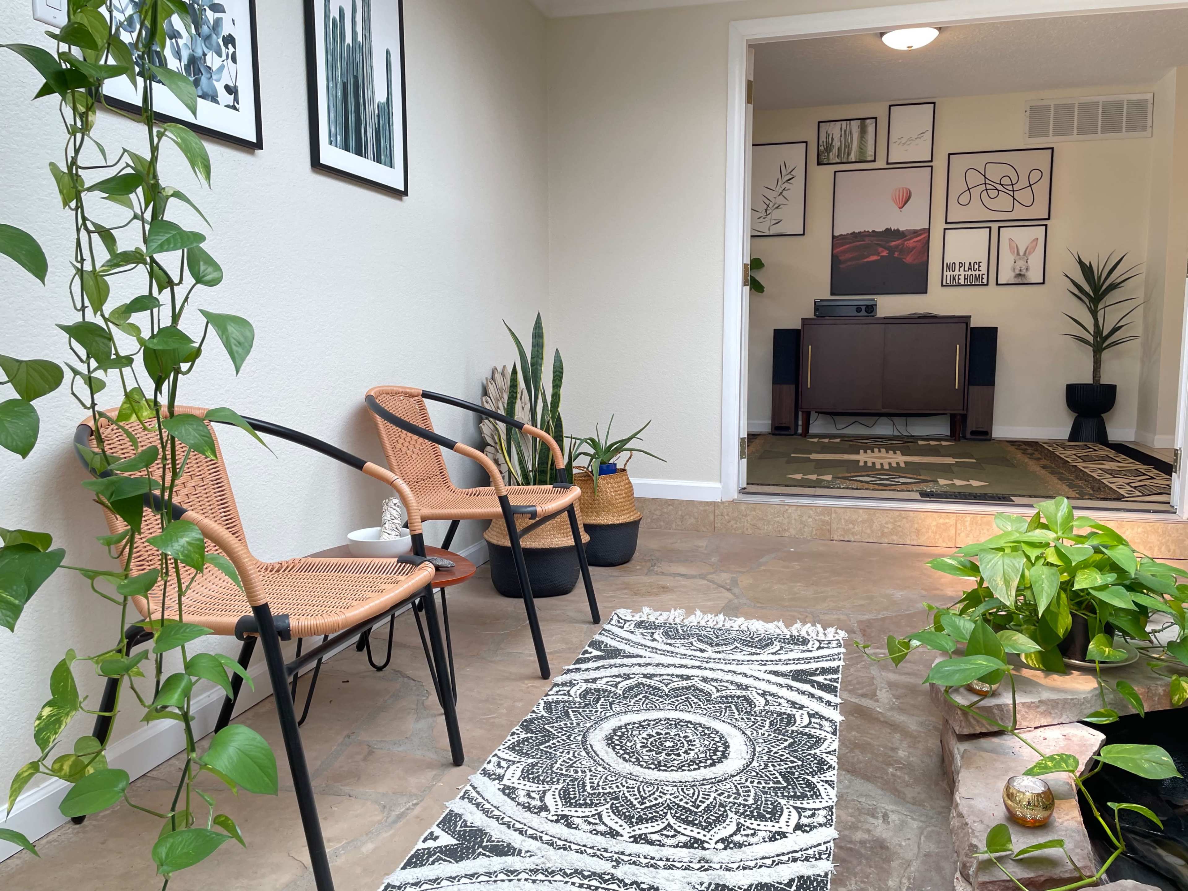 A cozy entryway features two woven chairs, a decorative rug, and potted plants near a staircase leading to a room with a media console.