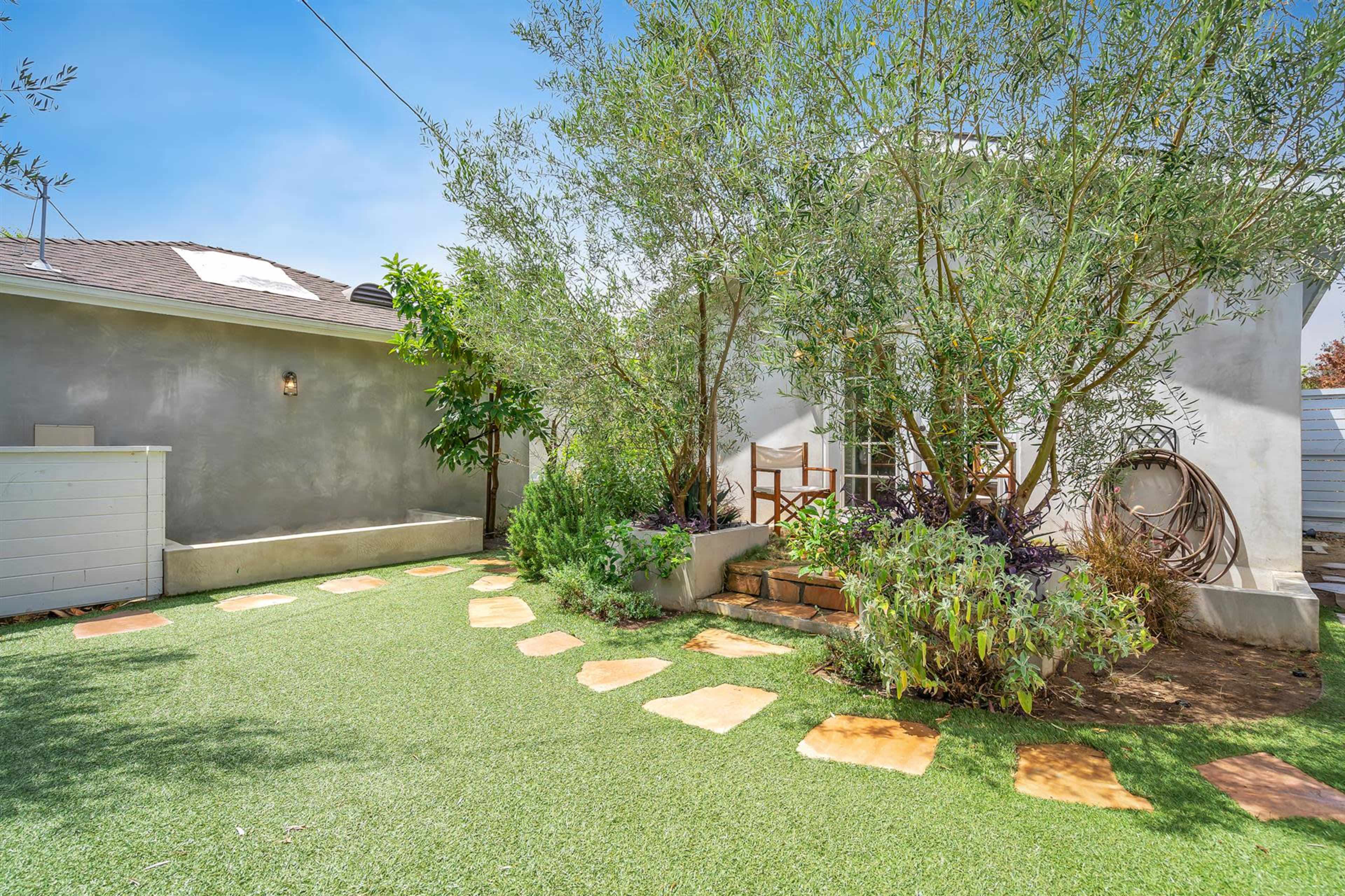 The image shows a small, landscaped yard with a path of stone slabs leading through greenery to a white building.