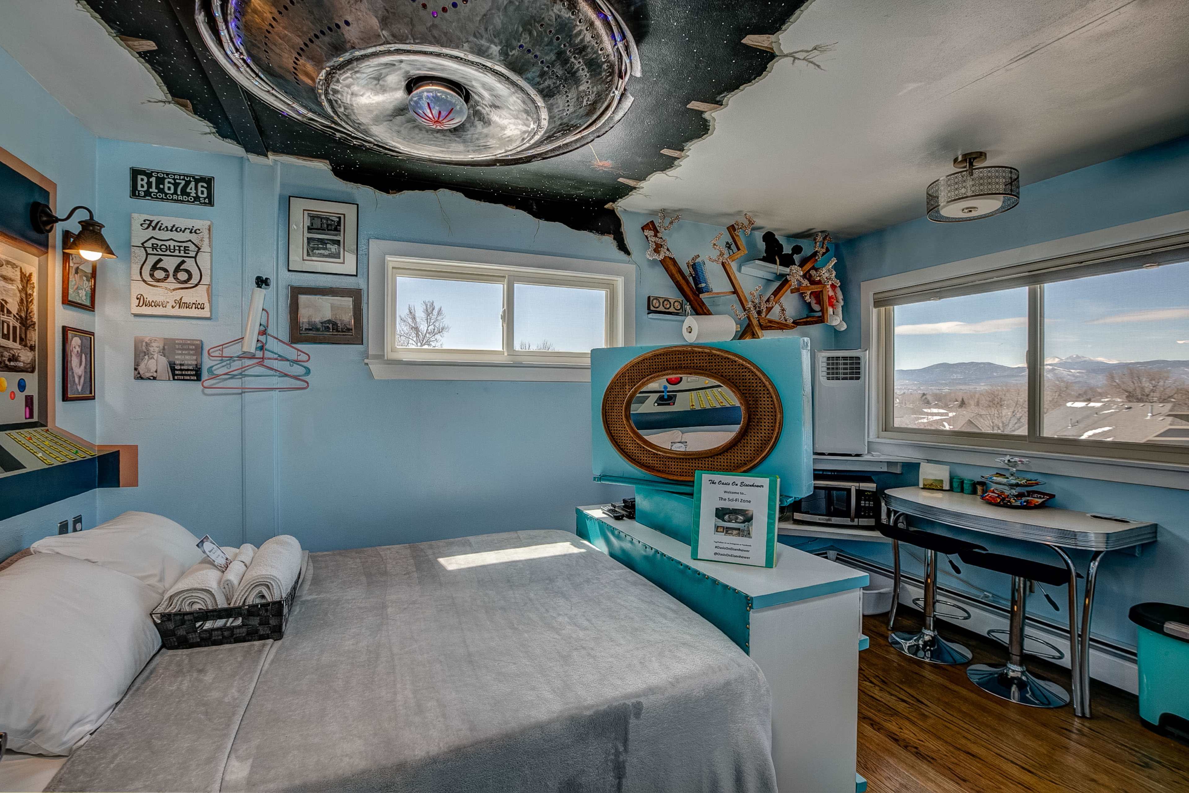 A bedroom features a mounted metallic disc light fixture on a painted ceiling, with a bed, blue walls adorned with various memorabilia, and a small dining table near a window overlooking mountains.