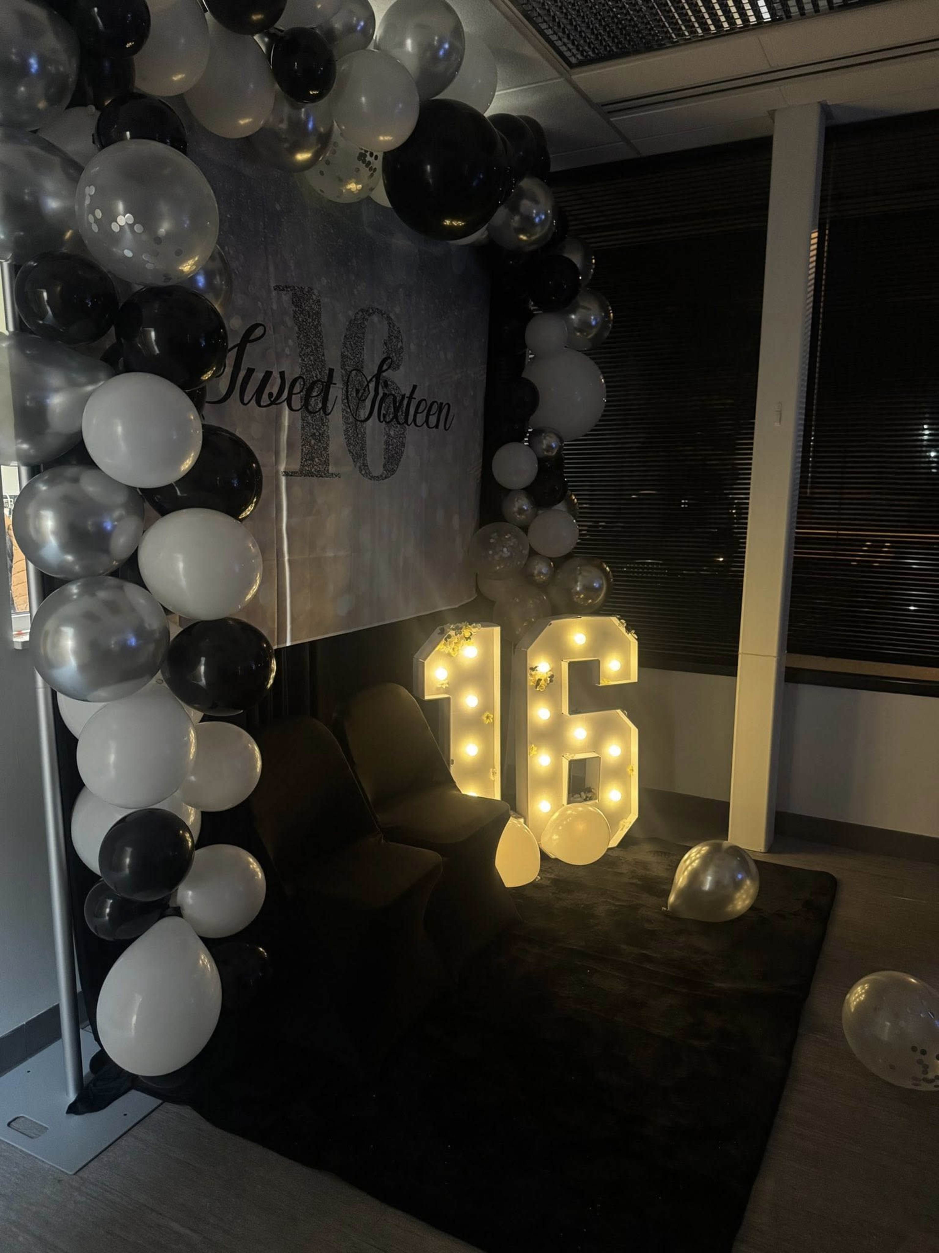 The image shows a decorated corner for a Sweet Sixteen celebration, featuring a backdrop with the text "Sweet Sixteen," large illuminated numbers "16," and an arch made of black, white, and silver balloons.