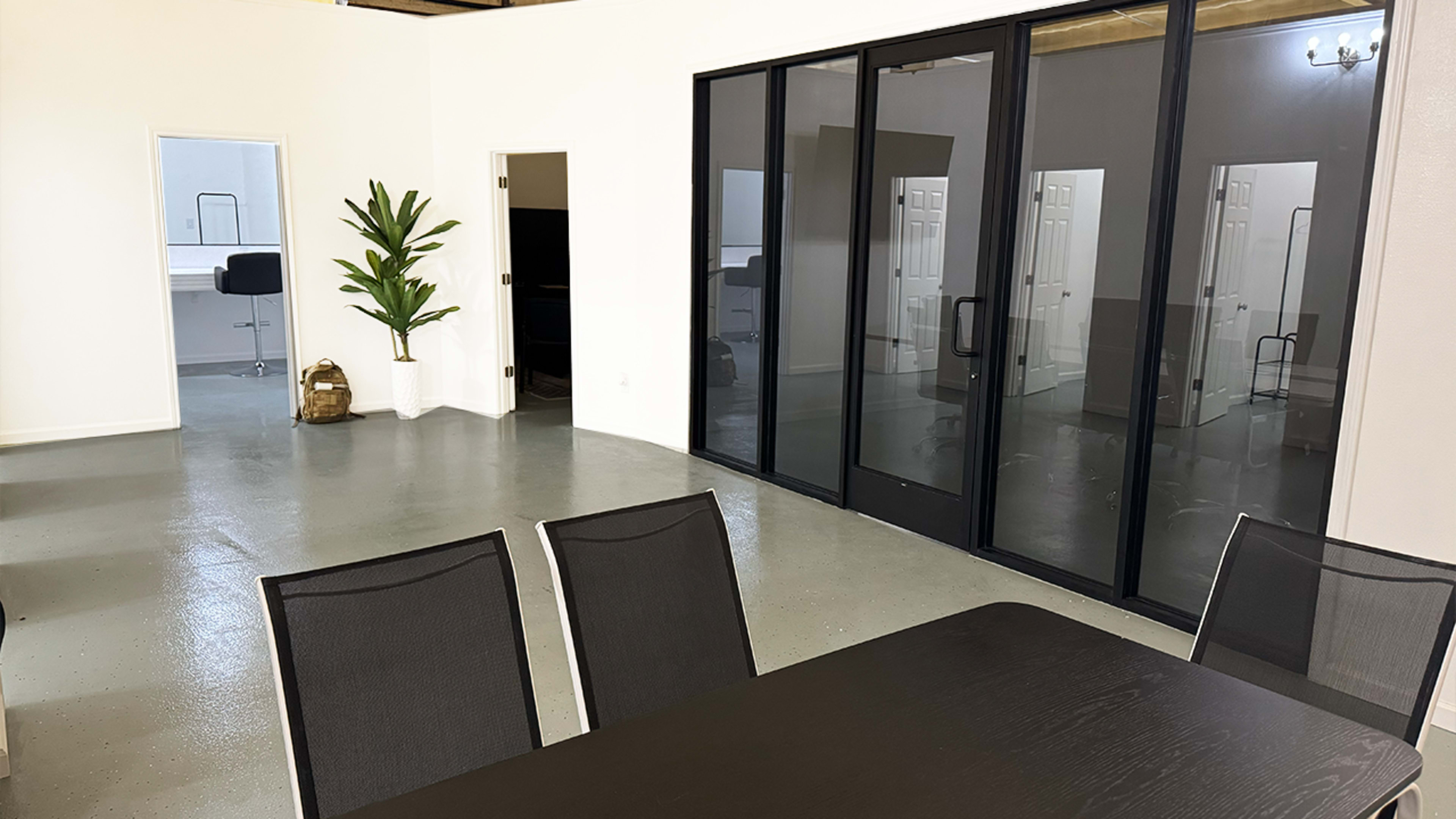 The image shows a spacious, modern office interior featuring a large table with chairs, a potted plant, and glass doors leading to a separate room.