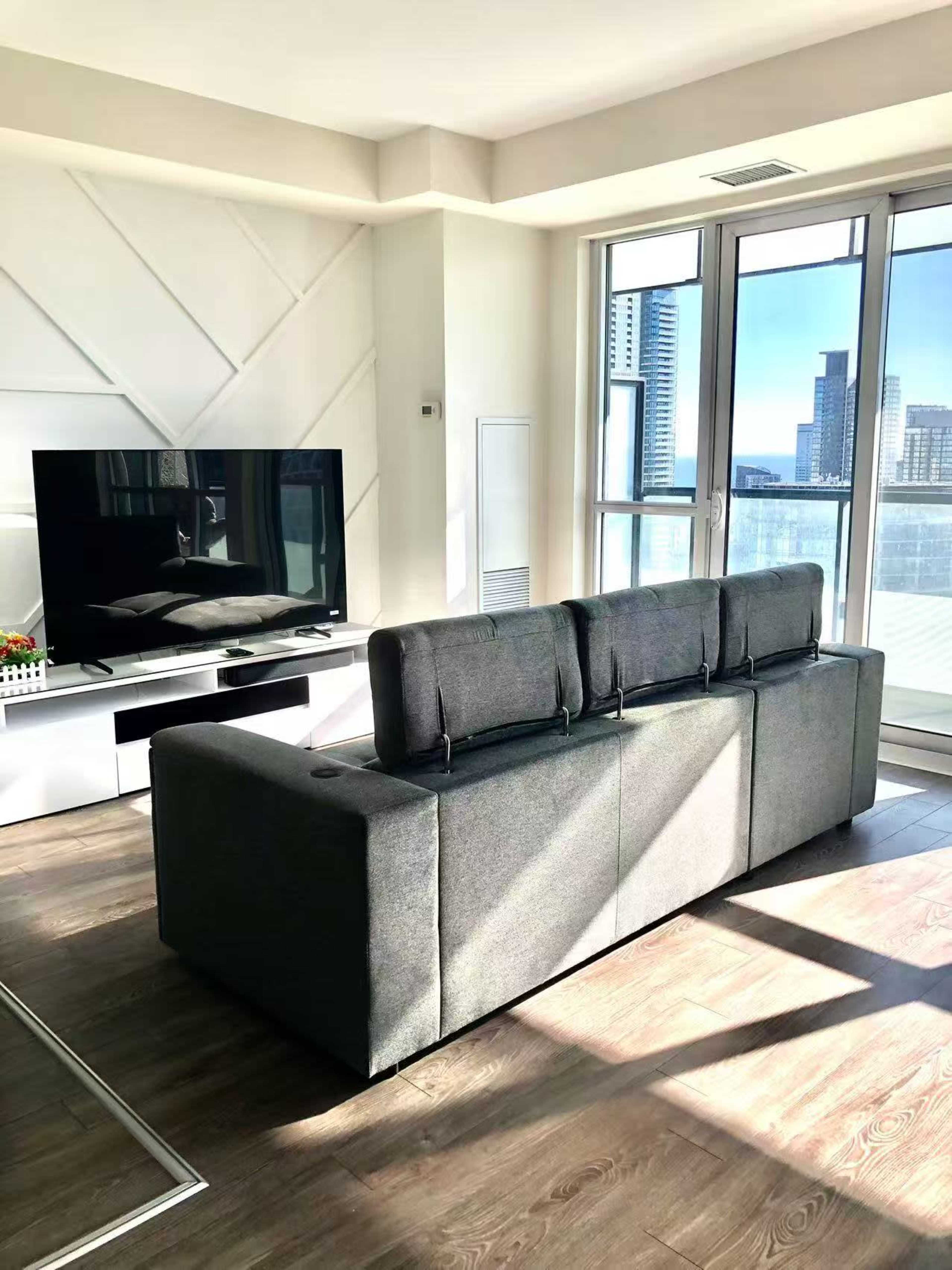 A modern living room features a gray sectional sofa facing a large flat-screen TV, with large windows allowing natural light to enter the space.