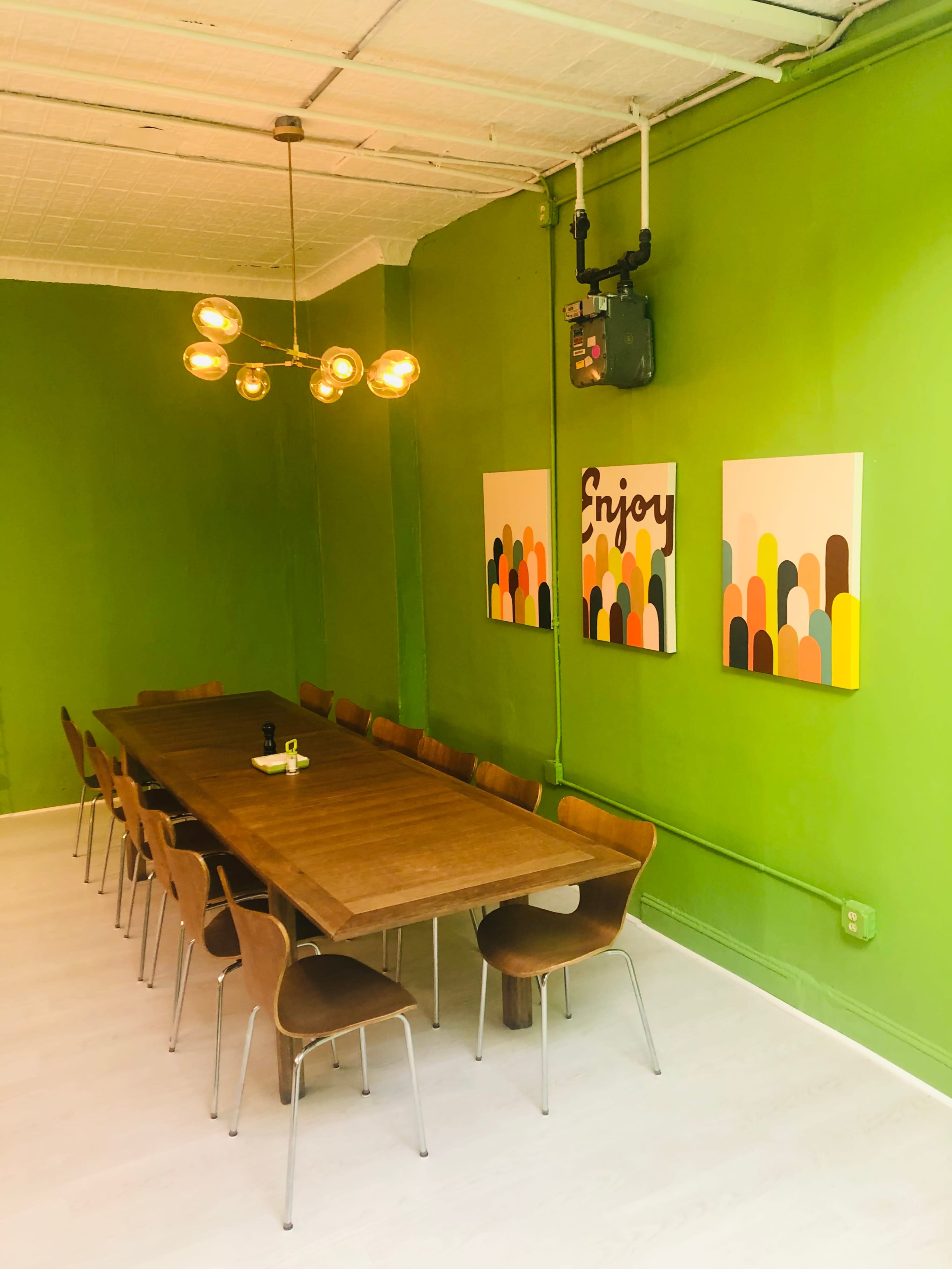 A long wooden table with chairs is positioned in a green-walled meeting room featuring two colorful abstract art pieces and a light fixture above.