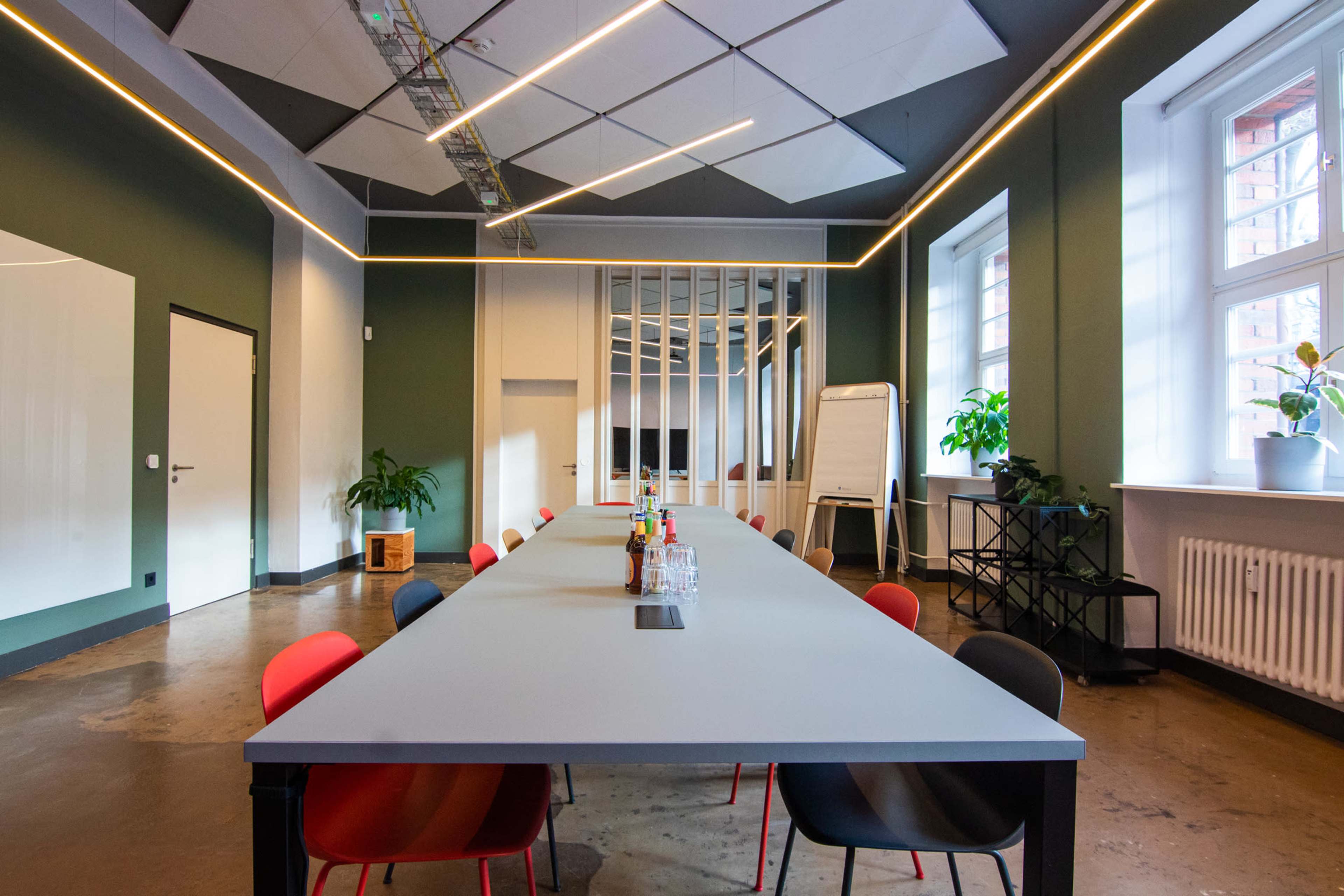 A long conference table is set in a modern meeting room with green walls, large windows, and a variety of colorful chairs.